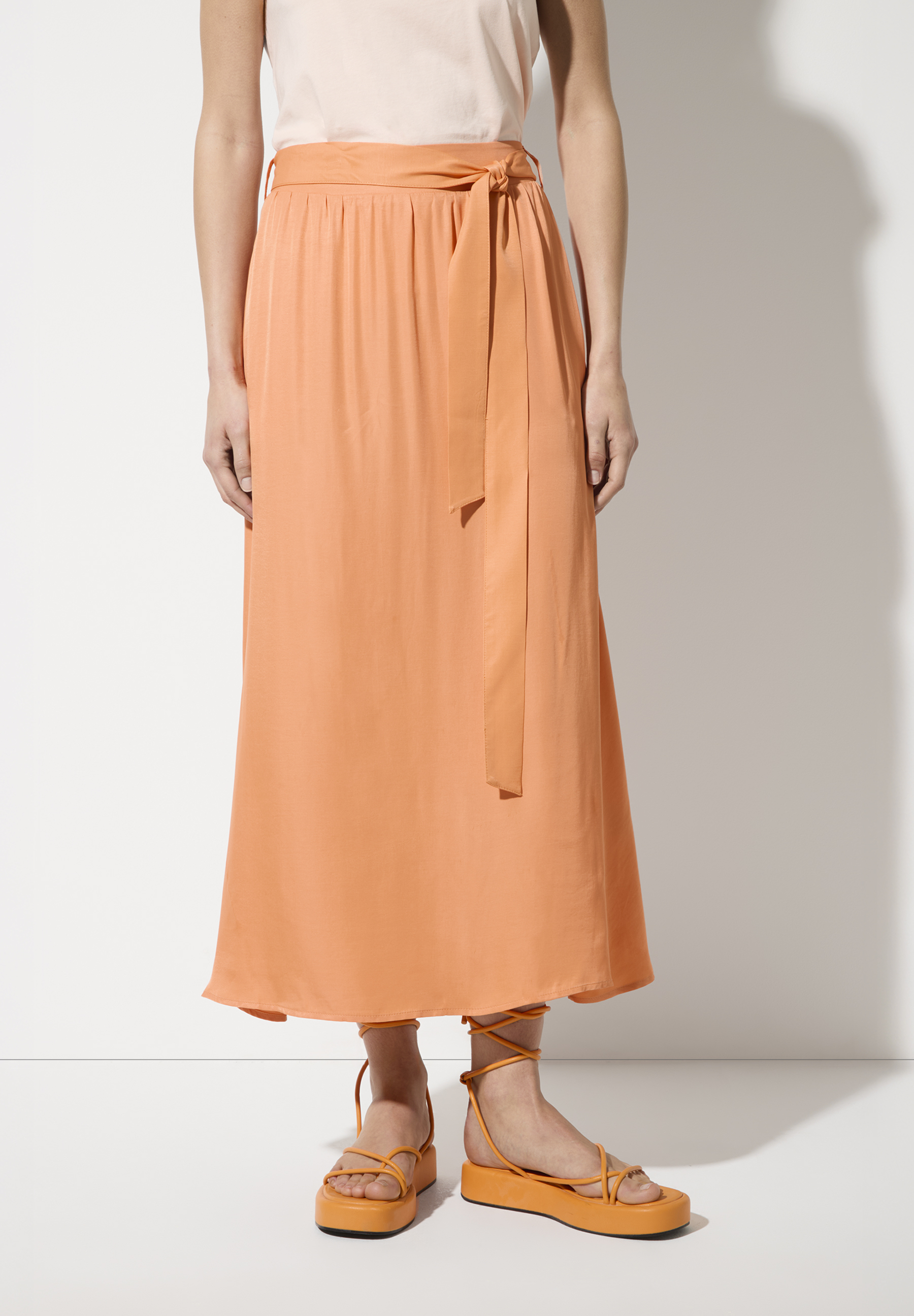 Ein Model trägt einen langen, orangefarbenen MORE & MORE Rock mit Bindegürtel.