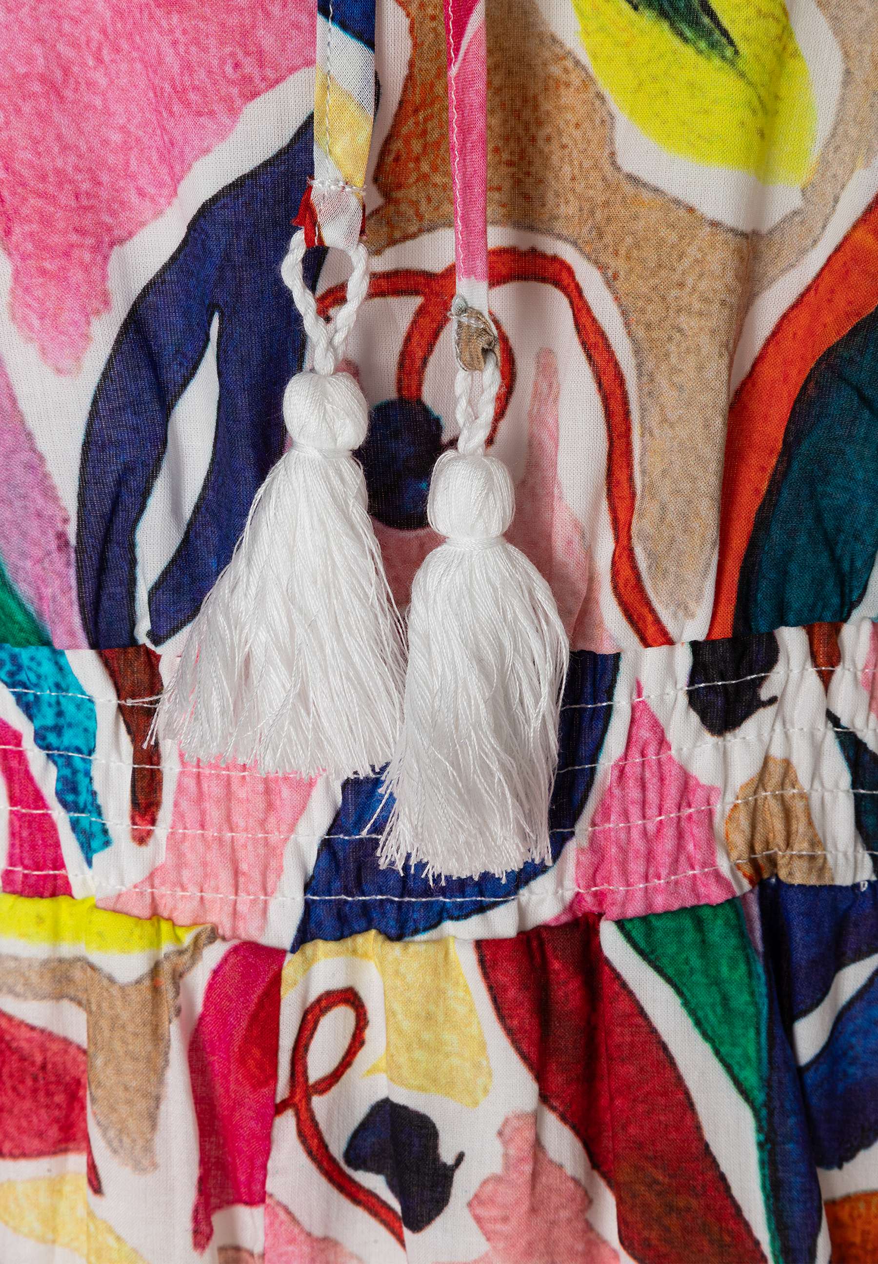 Detailansicht eines MORE & MORE Oberteils aus leichtem Voile-Material mit bunten Mustern und weißen Quasten. Detailansicht eines MORE & MORE Oberteils aus leichtem Voile-Material mit bunten Mustern und weißen Quasten.