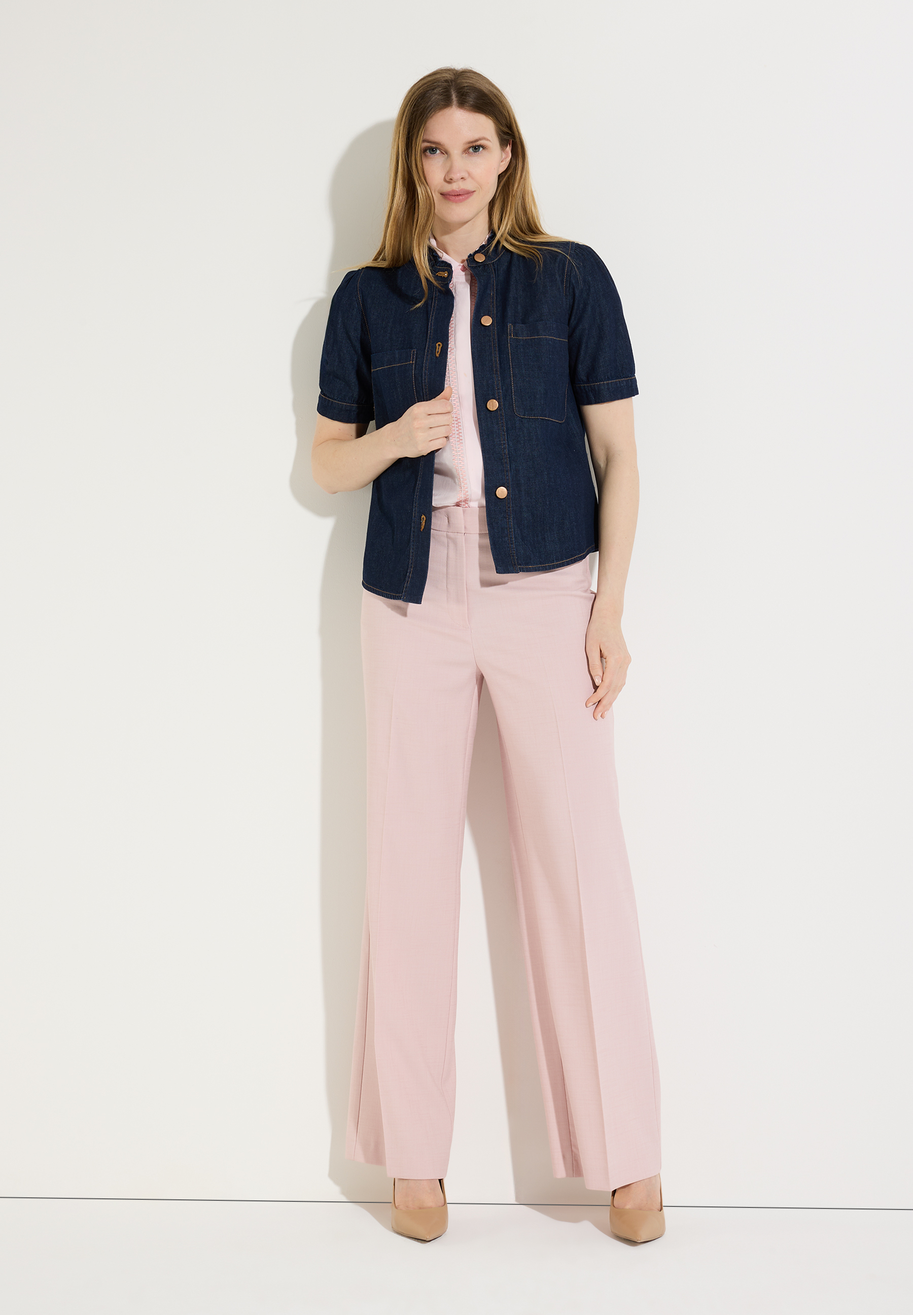 Eine Frau in MORE & MORE Bluse, hellrosa Hose und Schuhen vor einer weißen Wand. Eine Frau in MORE & MORE Bluse, hellrosa Hose und Schuhen vor einer weißen Wand.