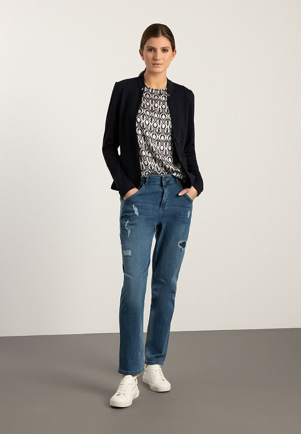 Eine Frau in MORE & MORE Jeans mit Blazer und Sneakern vor hellem Hintergrund. Eine Frau in MORE & MORE Jeans mit Blazer und Sneakern vor hellem Hintergrund.