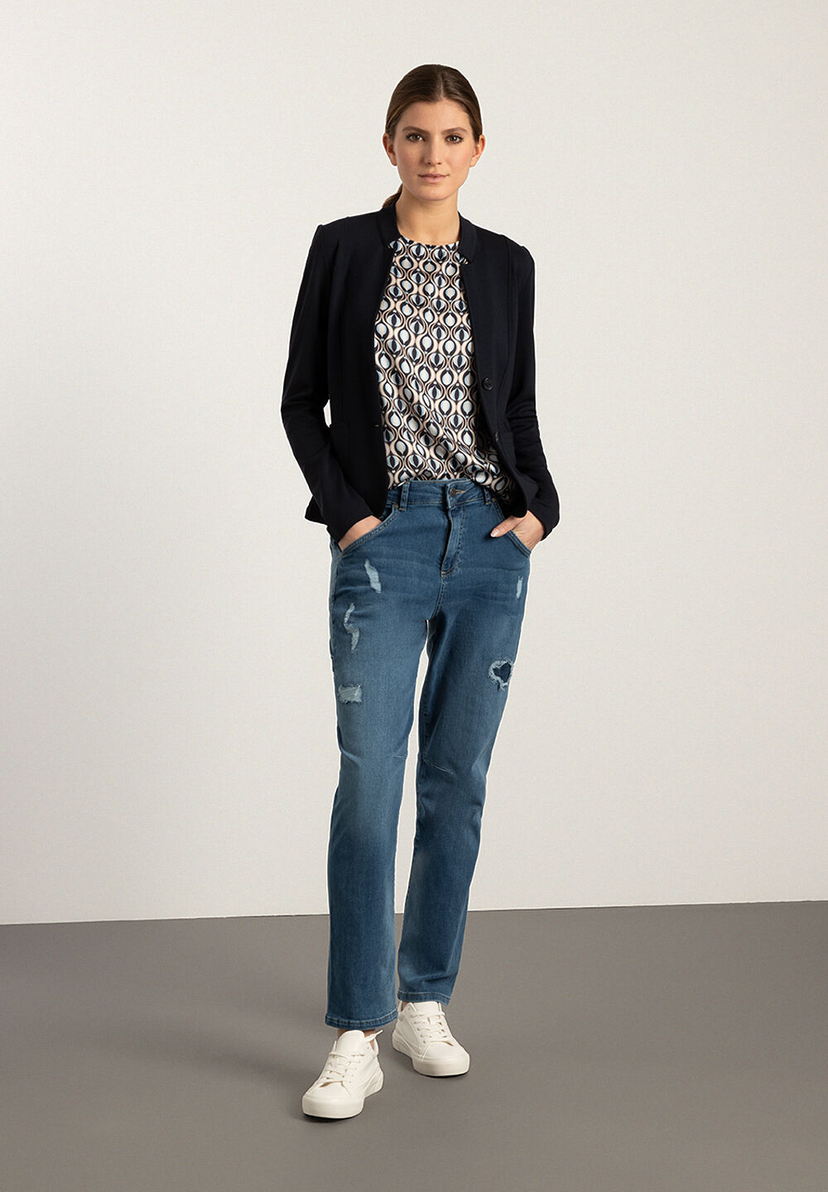 Eine Frau in MORE & MORE Jeans mit Blazer und Sneakern vor hellem Hintergrund. Eine Frau in MORE & MORE Jeans mit Blazer und Sneakern vor hellem Hintergrund.