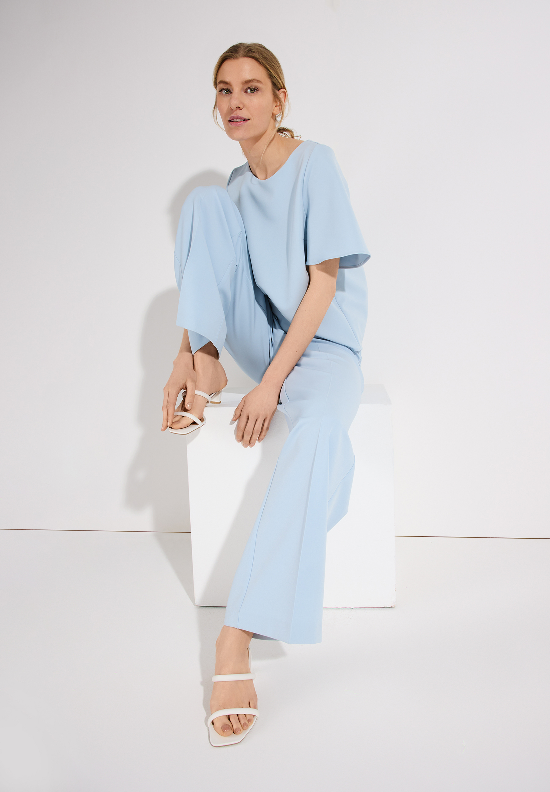 Eine Frau in einem hellblauen MORE & MORE Oberteil und Hose sitzt auf einem weißen Würfel. Eine Frau in einem hellblauen MORE & MORE Oberteil und Hose sitzt auf einem weißen Würfel.