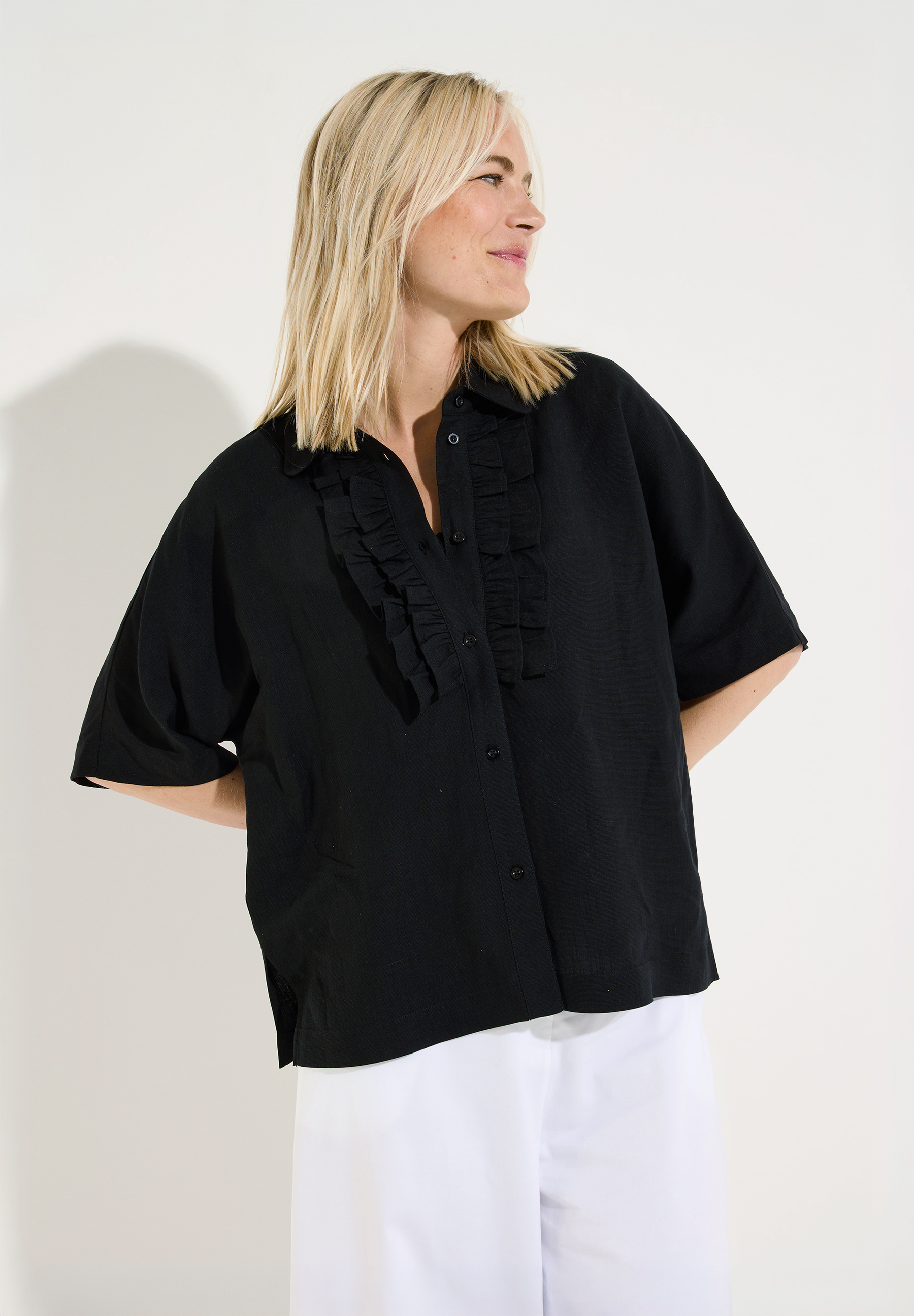 MORE & MORE schwarze Bluse mit Rüschen-Detail, locker fallend und kurzen Ärmeln, kombiniert mit weißer Hose.