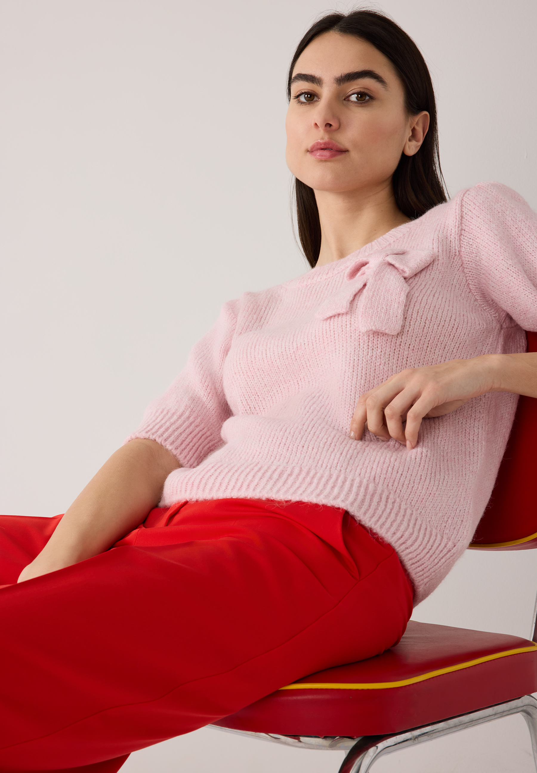 Eine Frau sitzt in einem MORE & MORE Strickoberteil/Pullover und roter Hose. Eine Frau sitzt in einem MORE & MORE Strickoberteil/Pullover und roter Hose.