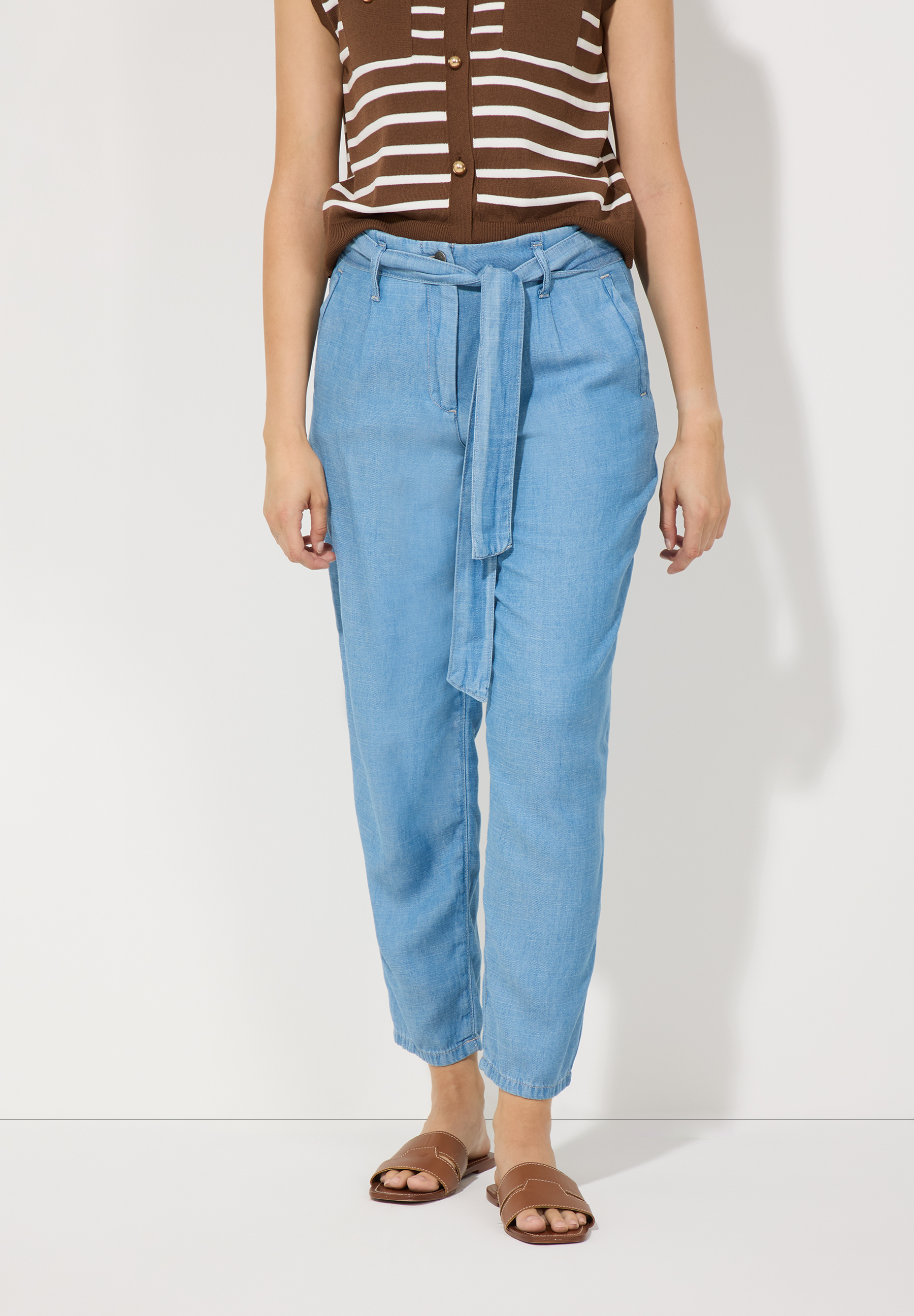 Eine Person trägt blaue Jeans der Marke MORE & MORE mit Bindegürtel und braune Sandalen vor einem weißen Hintergrund.