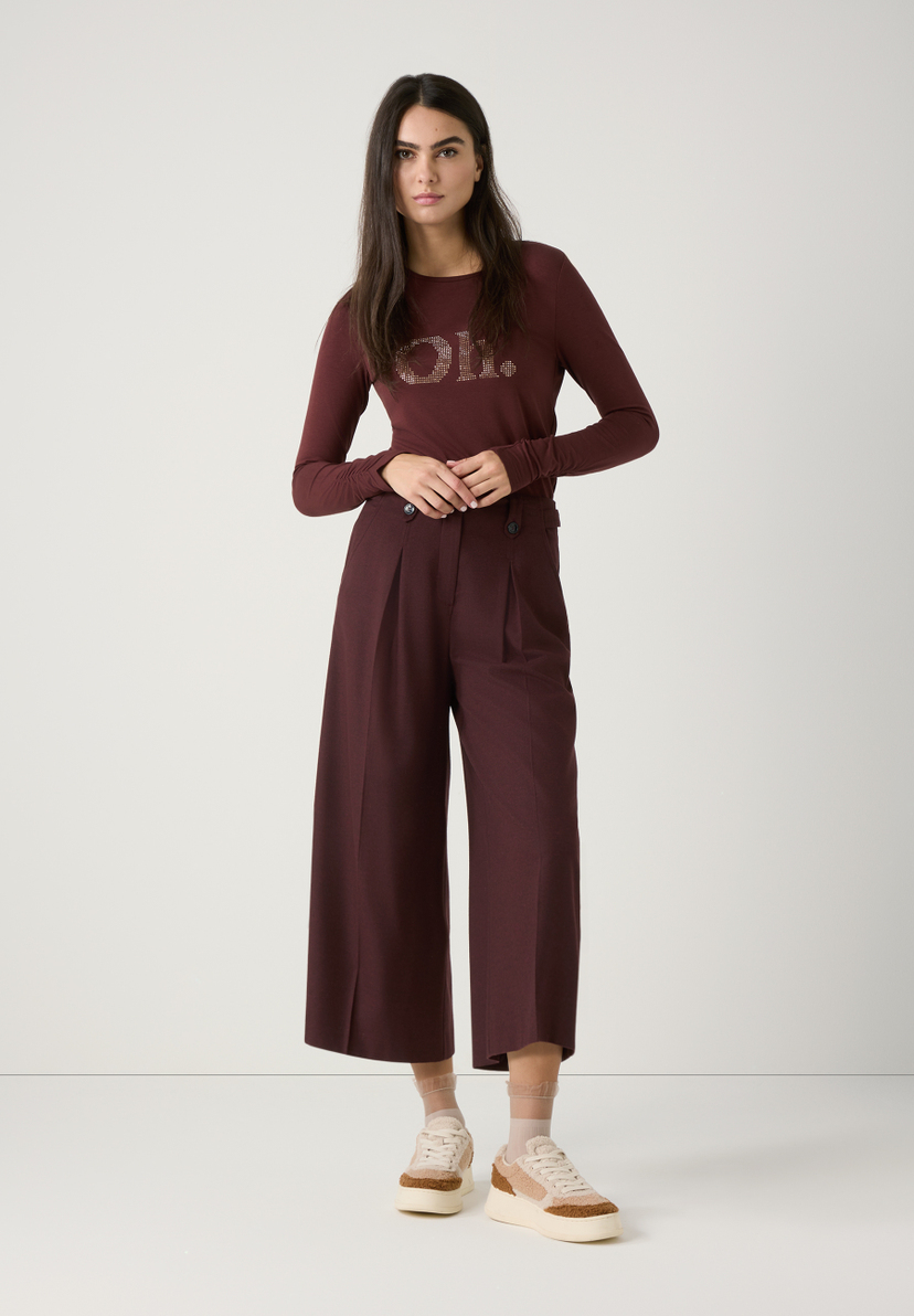 Eine Frau trägt ein Outfit von MORE & MORE: Ein weinrotes T-Shirt und eine weite Hose mit Sneakern. Eine Frau trägt ein Outfit von MORE & MORE: Ein weinrotes T-Shirt und eine weite Hose mit Sneakern.