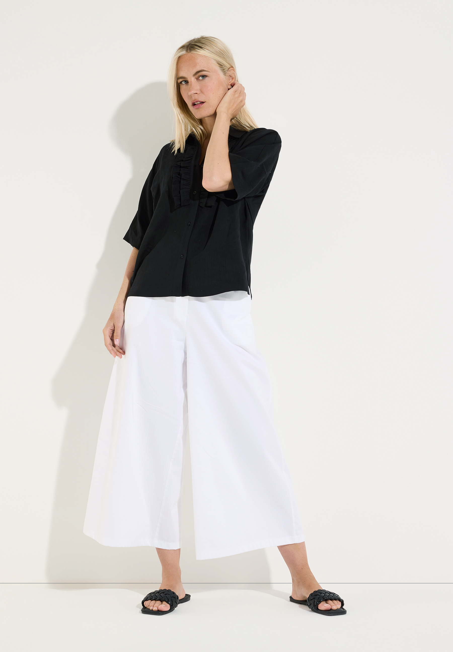 MORE & MORE Bluse: Eine blonde Frau trägt eine schwarze, kurze Bluse mit Rüschen am Ausschnitt und eine weite weiße Hose. MORE & MORE Bluse: Eine blonde Frau trägt eine schwarze, kurze Bluse mit Rüschen am Ausschnitt und eine weite weiße Hose.