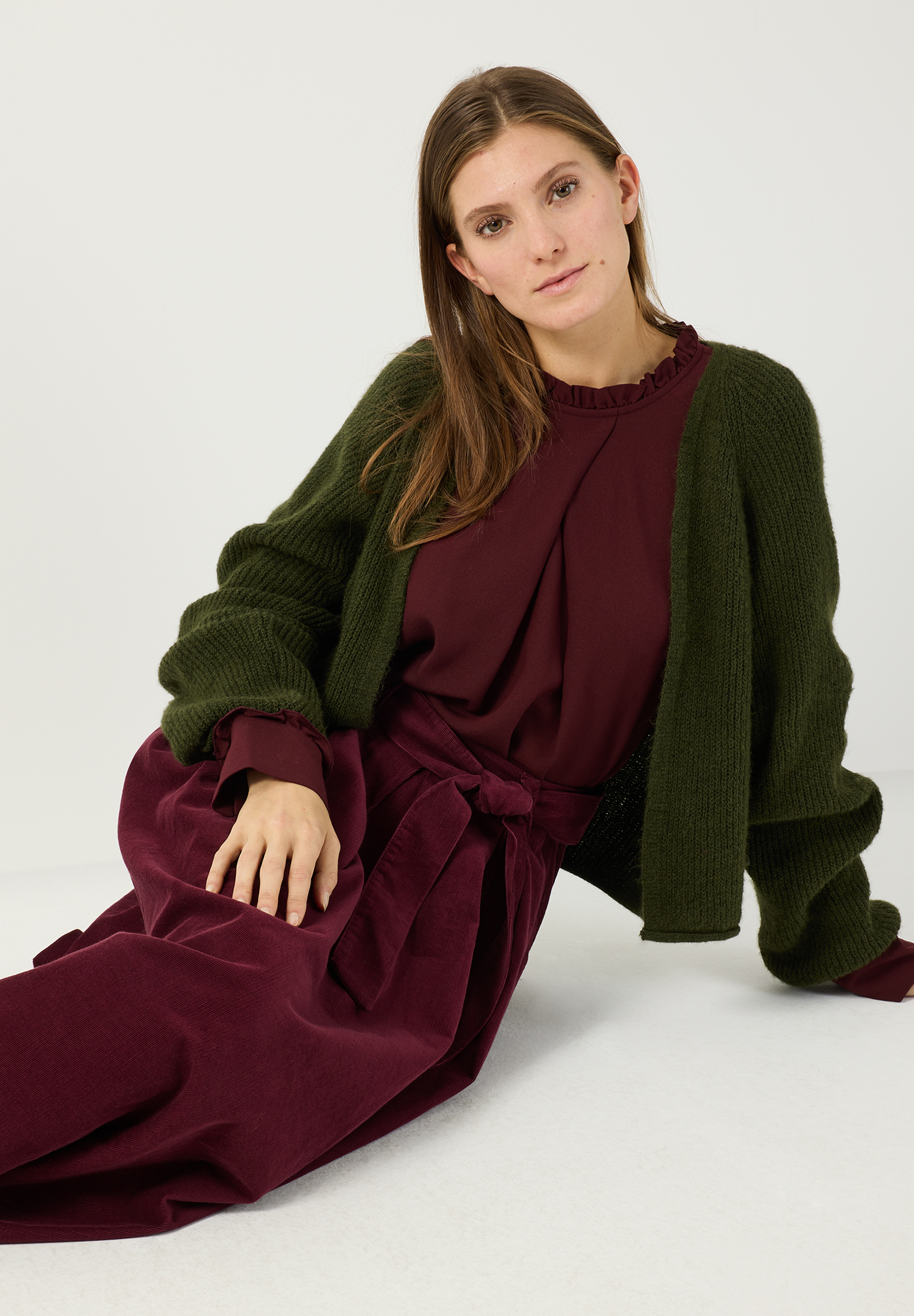 MORE & MORE Pullover: Eine junge Frau im bordeauxroten Outfit mit gerüschtem Kragen und khakifarbenem Strickpullover MORE & MORE Pullover: Eine junge Frau im bordeauxroten Outfit mit gerüschtem Kragen und khakifarbenem Strickpullover