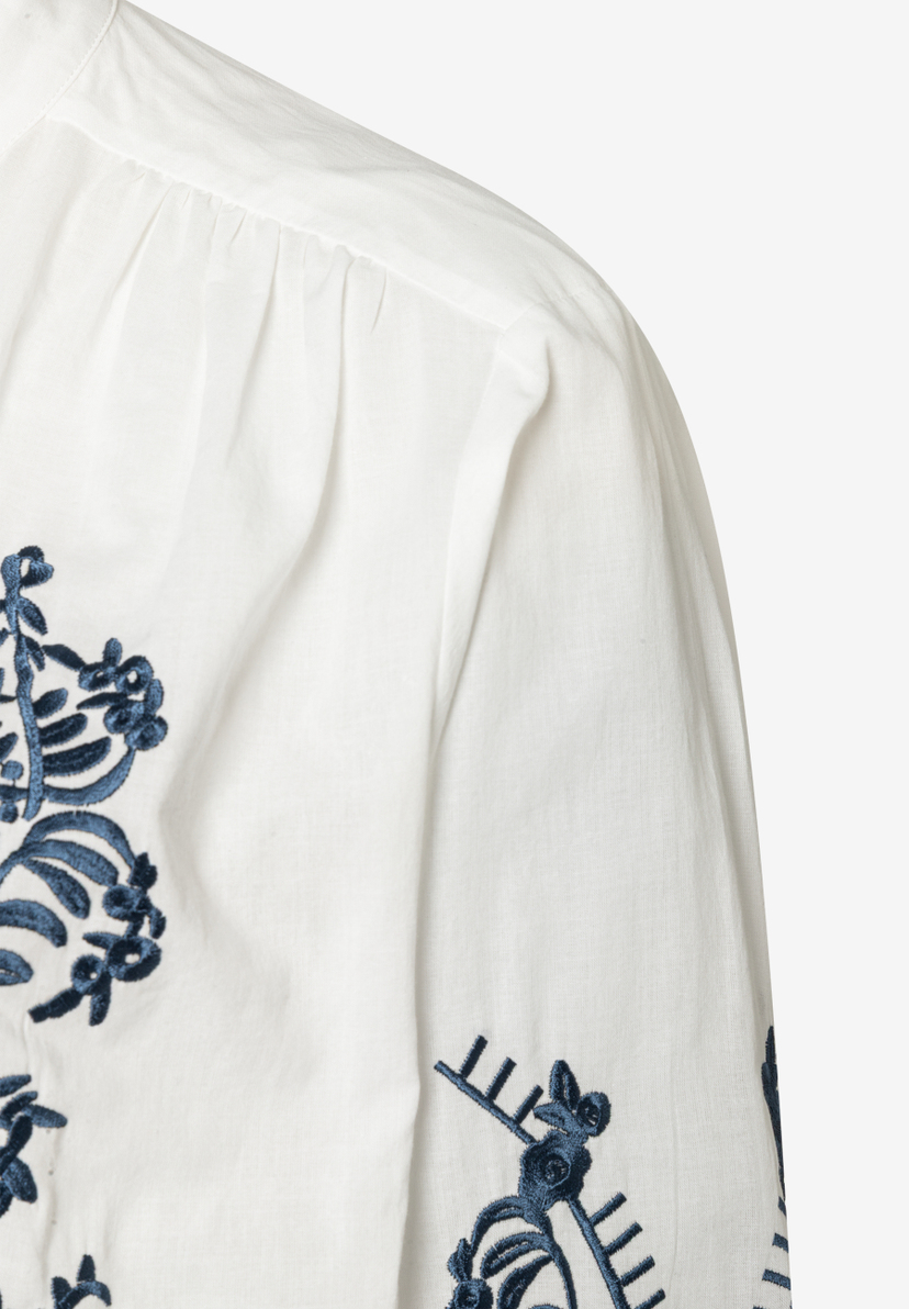 Detailansicht einer weißen MORE & MORE Bluse mit blauen Stickereien an Ärmel und Schulter. Detailansicht einer weißen MORE & MORE Bluse mit blauen Stickereien an Ärmel und Schulter.