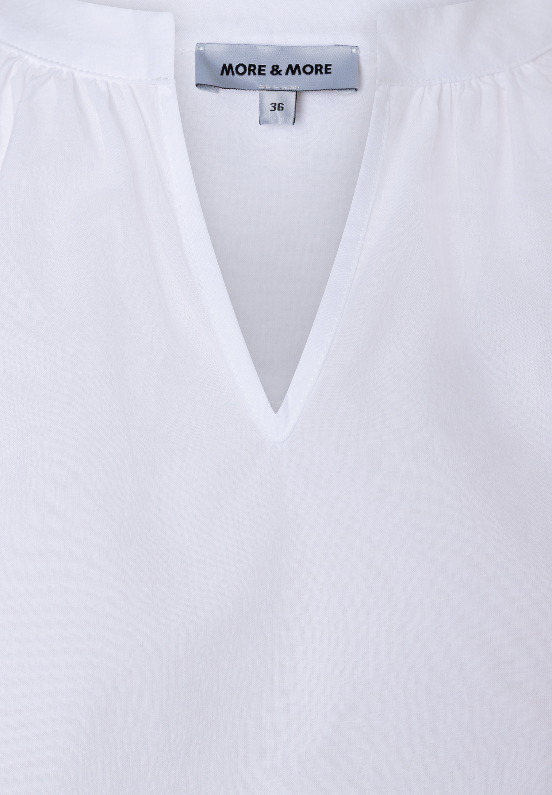 Detailaufnahme einer weißen MORE & MORE Bluse aus Baumwolle mit V-Ausschnitt und Größenetikett. Detailaufnahme einer weißen MORE & MORE Bluse aus Baumwolle mit V-Ausschnitt und Größenetikett.