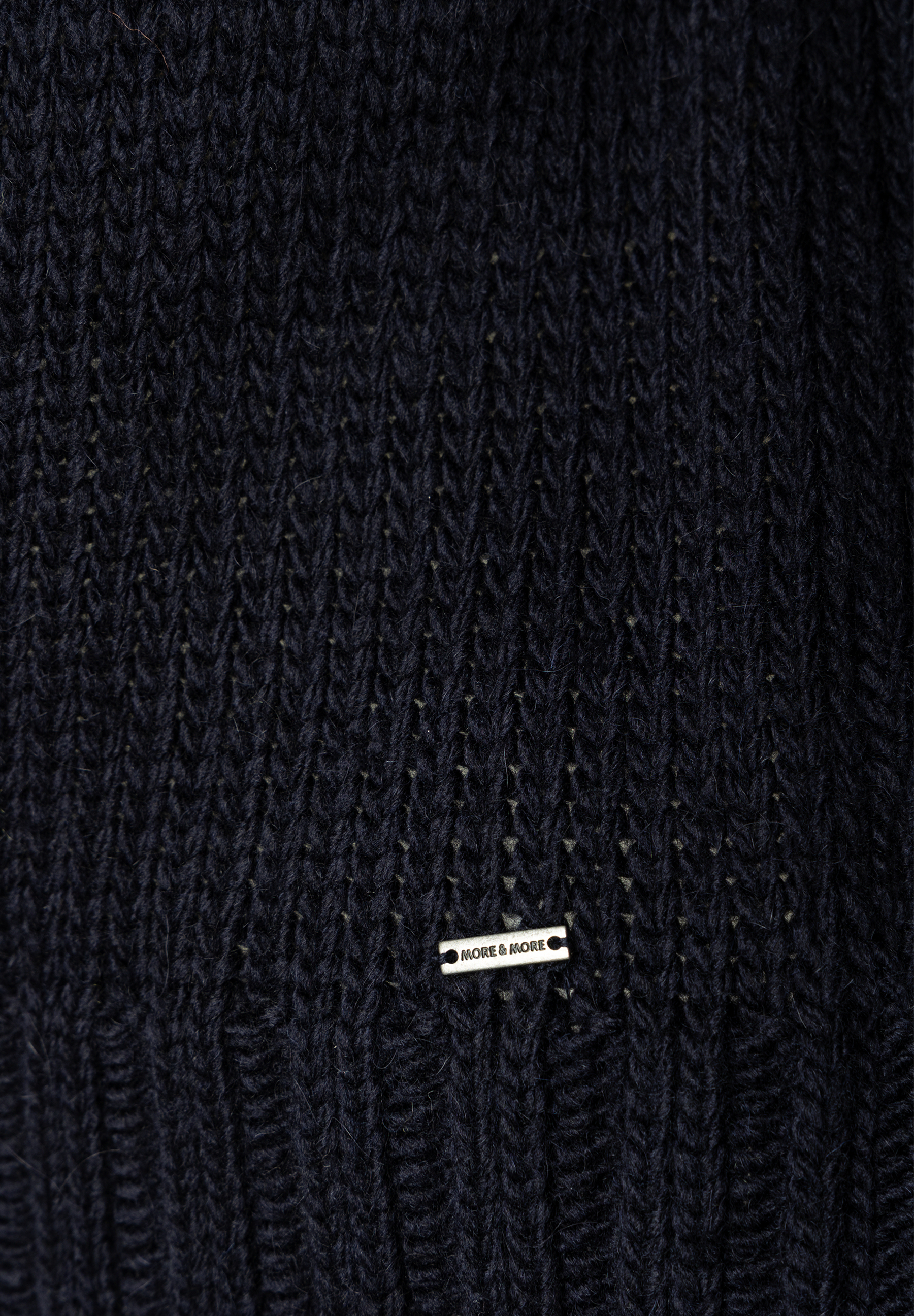 MORE & MORE Pullover: Detailaufnahme des dunkelblauen Strickmusters mit eingearbeitetem silbernen Markenlogo. MORE & MORE Pullover: Detailaufnahme des dunkelblauen Strickmusters mit eingearbeitetem silbernen Markenlogo.