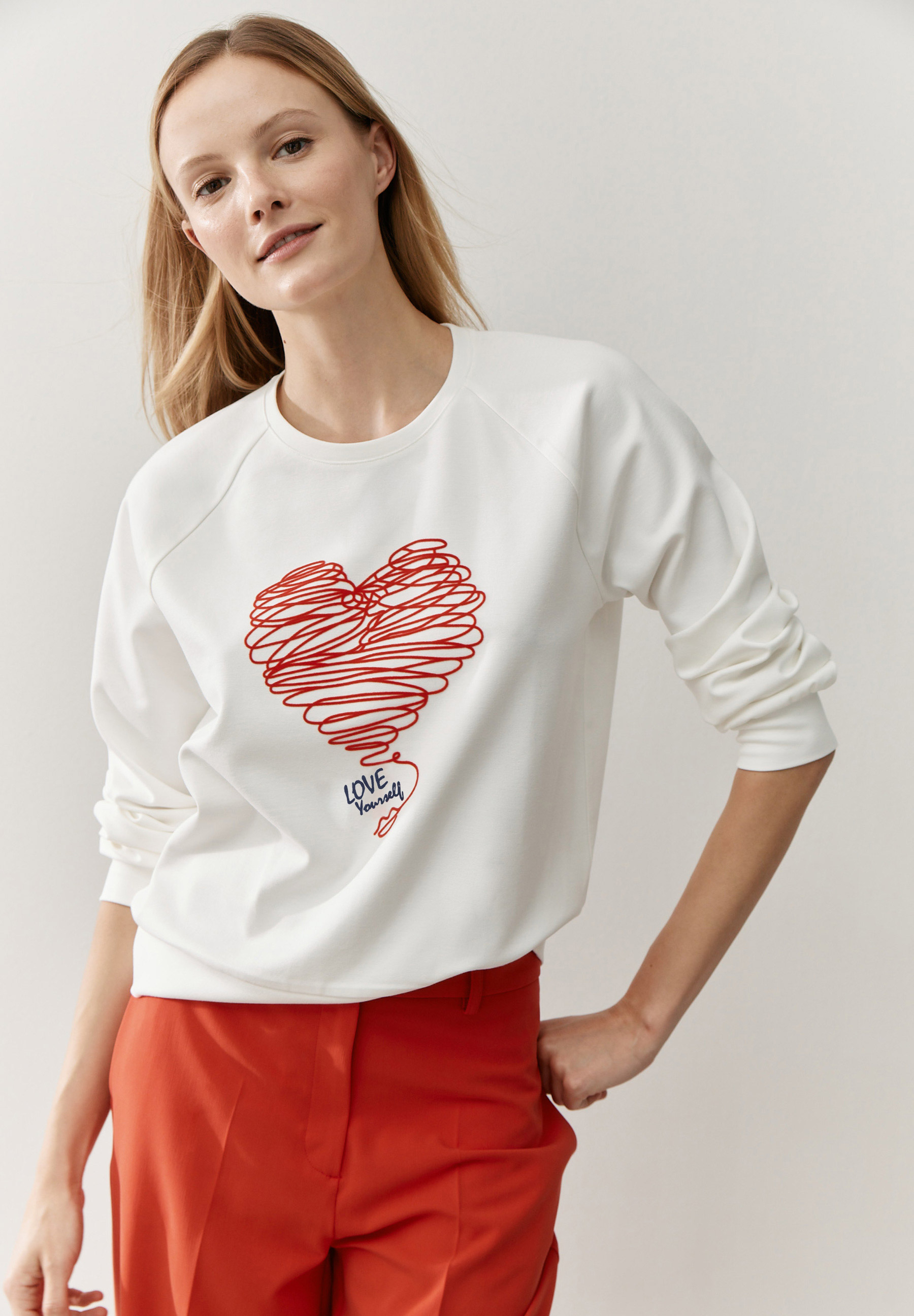 Eine Frau trägt ein MORE & MORE Sweatshirt mit einem roten Herz und roter Hose. Eine Frau trägt ein MORE & MORE Sweatshirt mit einem roten Herz und roter Hose.