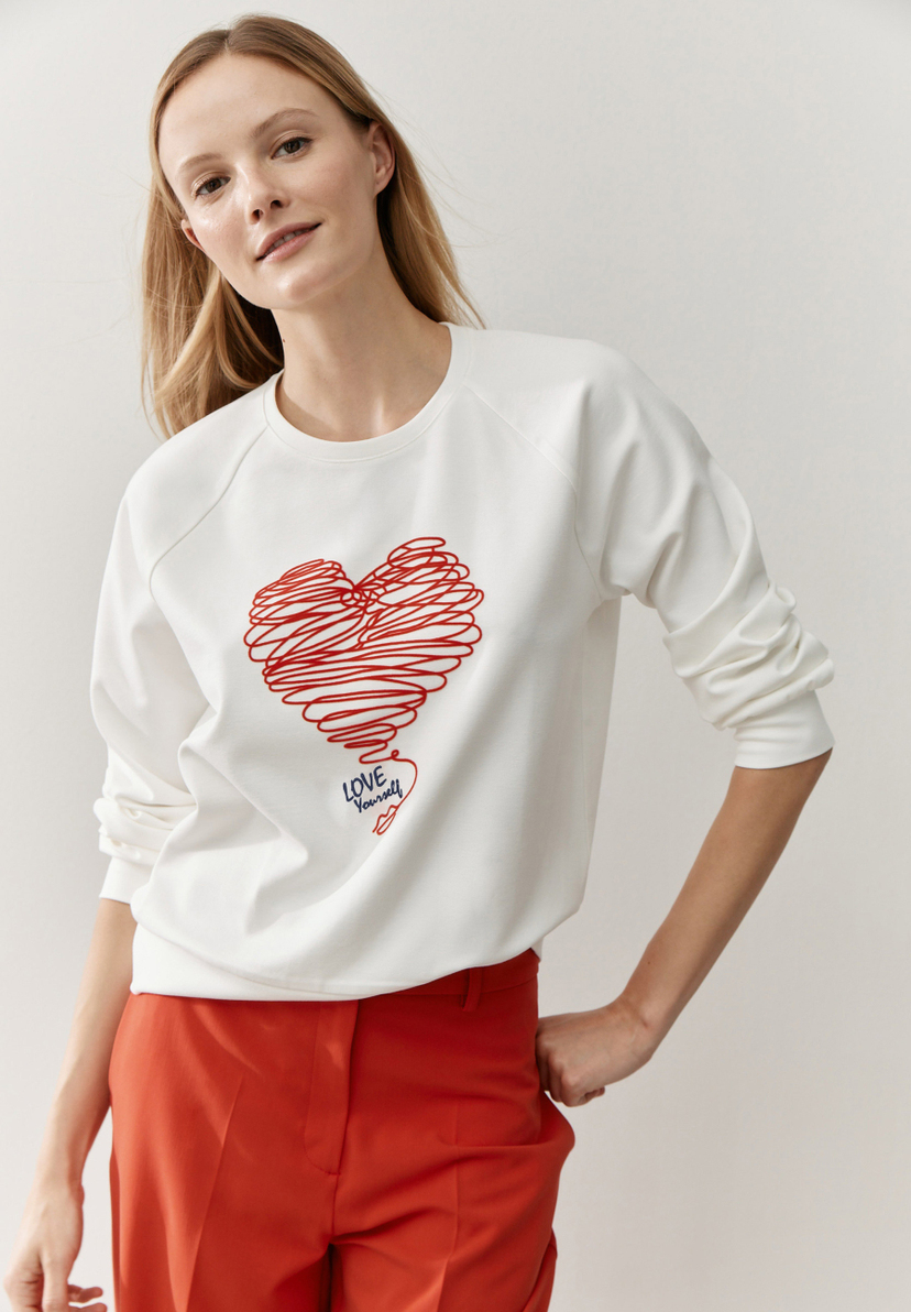 Eine Frau trägt ein MORE & MORE Sweatshirt mit einem roten Herz und roter Hose. Eine Frau trägt ein MORE & MORE Sweatshirt mit einem roten Herz und roter Hose.