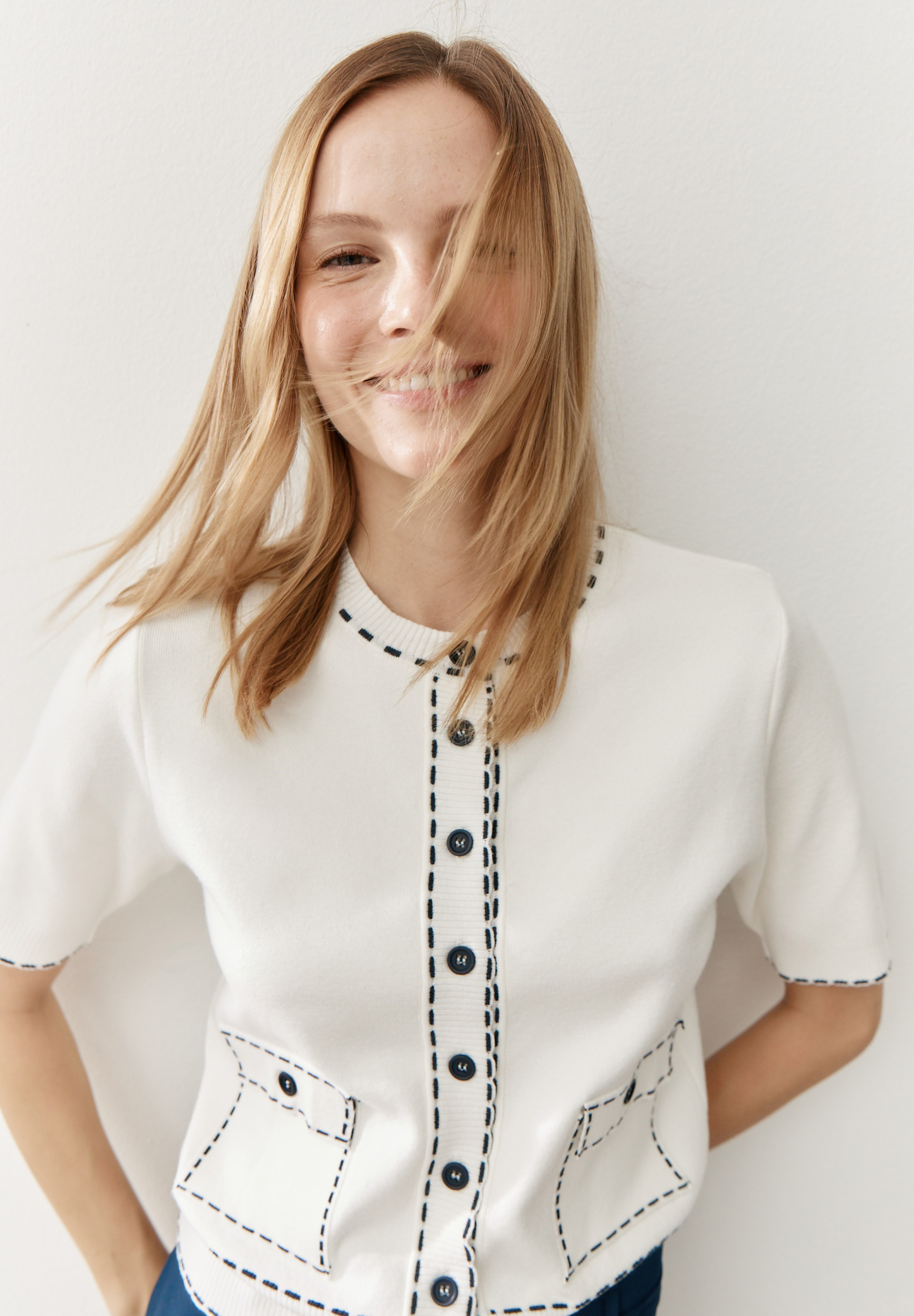 Eine lächelnde Frau mit blonden Haaren trägt einen weißen Strick-Cardigan von MORE & MORE mit Ziernähten.