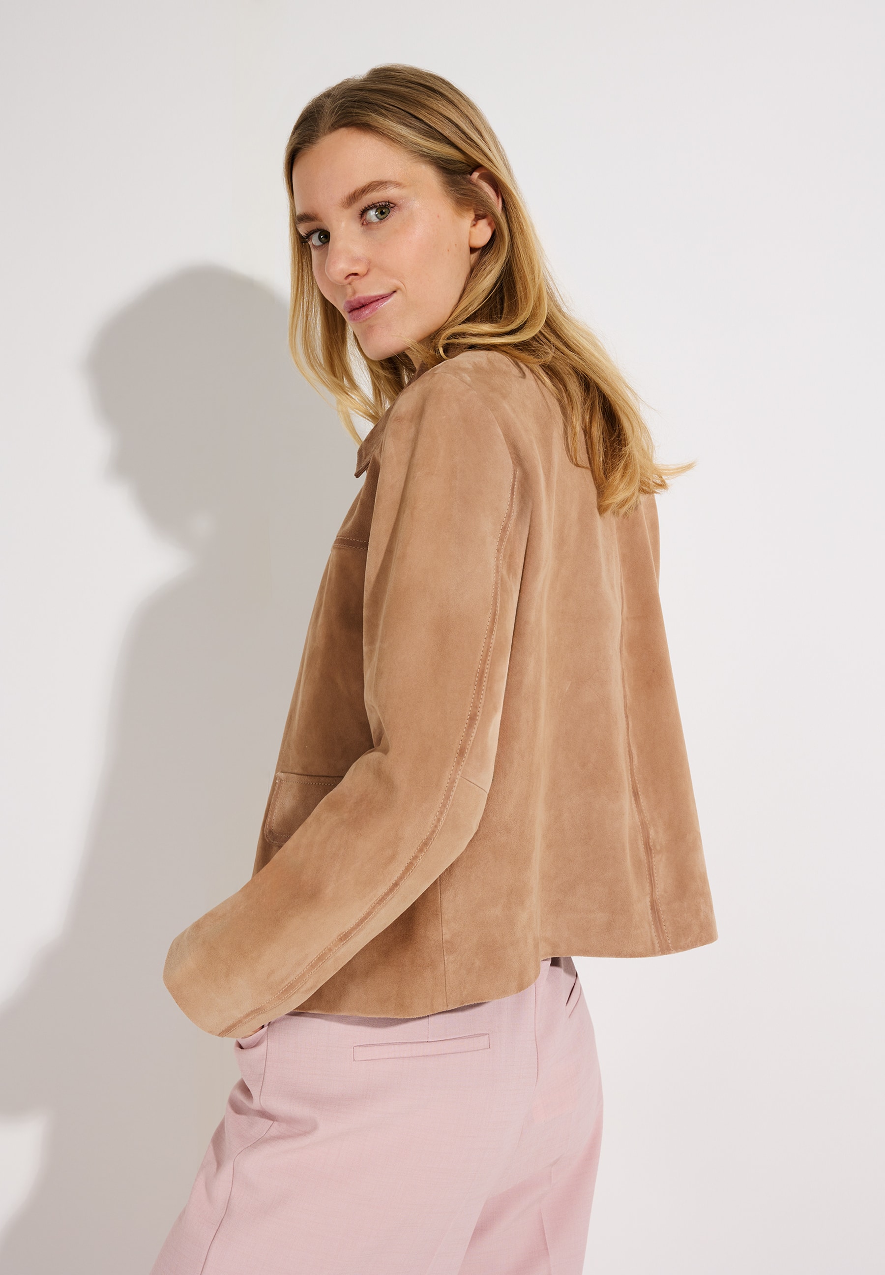 Eine Frau blickt über ihre Schulter. Sie trägt eine MORE & MORE Velourslederjacke und eine rosafarbene Hose. Eine Frau blickt über ihre Schulter. Sie trägt eine MORE & MORE Velourslederjacke und eine rosafarbene Hose.