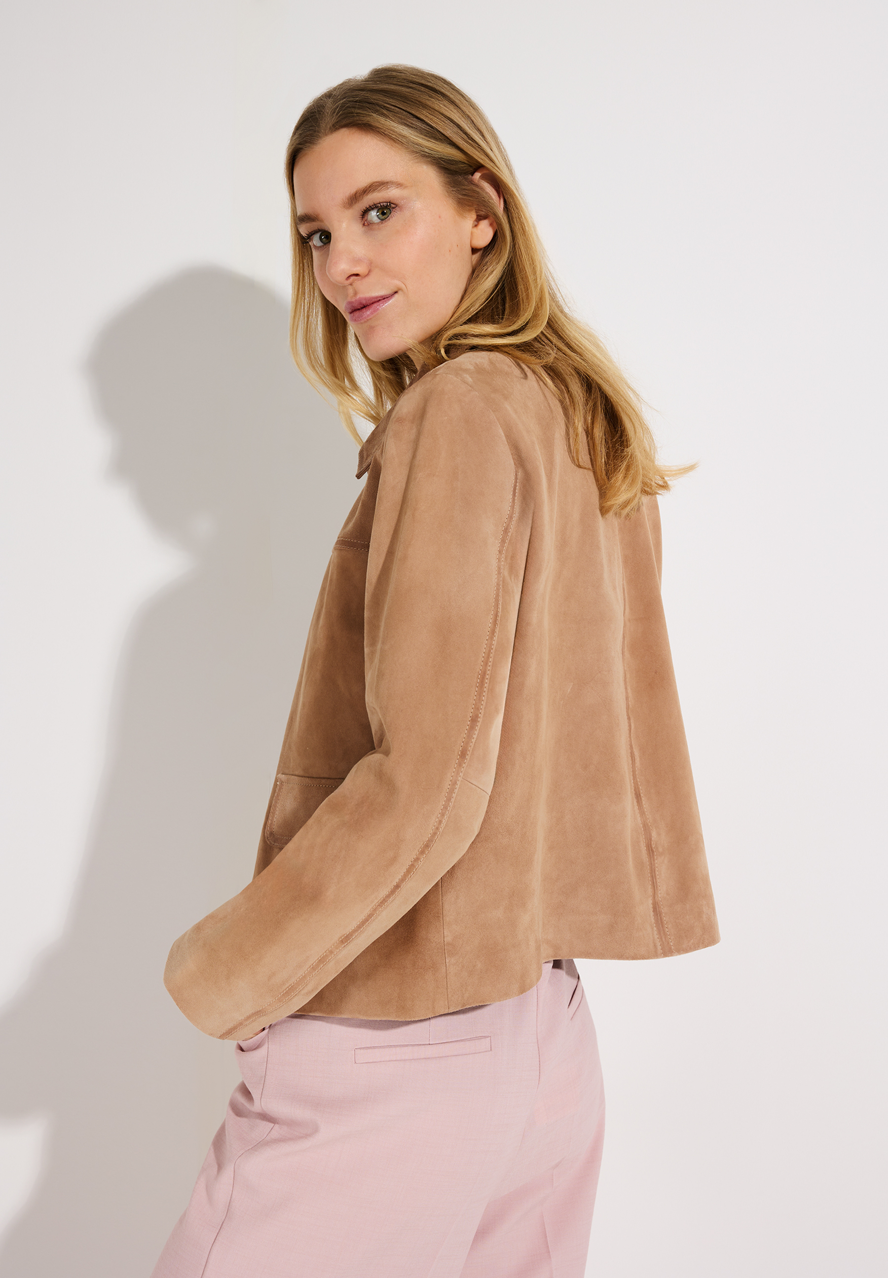 Eine Frau blickt über ihre Schulter. Sie trägt eine MORE & MORE Velourslederjacke und eine rosafarbene Hose. Eine Frau blickt über ihre Schulter. Sie trägt eine MORE & MORE Velourslederjacke und eine rosafarbene Hose.