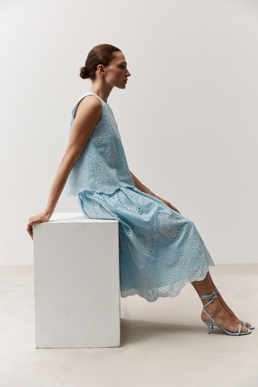 Eine Frau sitzt, gekleidet in ein blaues MORE & MORE-Spitzenoberteil und einen passenden Rock, dazu Sandalen.