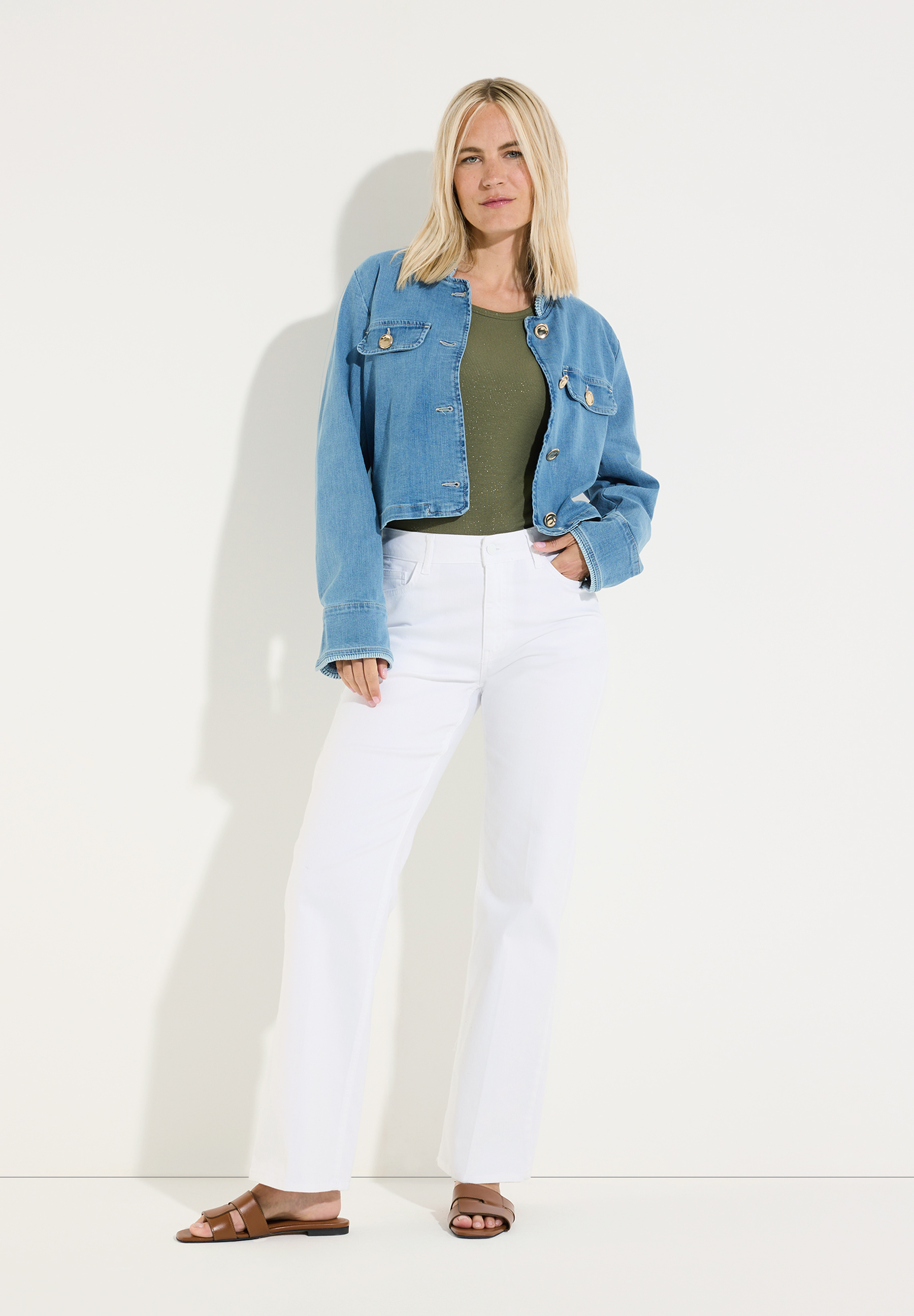 MORE & MORE Jeans: Blonde Frau in blauer Jeansjacke über grünem Top & weißer Hose. Trägt braune Sandalen, Hand in Hosentasche. MORE & MORE Jeans: Blonde Frau in blauer Jeansjacke über grünem Top & weißer Hose. Trägt braune Sandalen, Hand in Hosentasche.