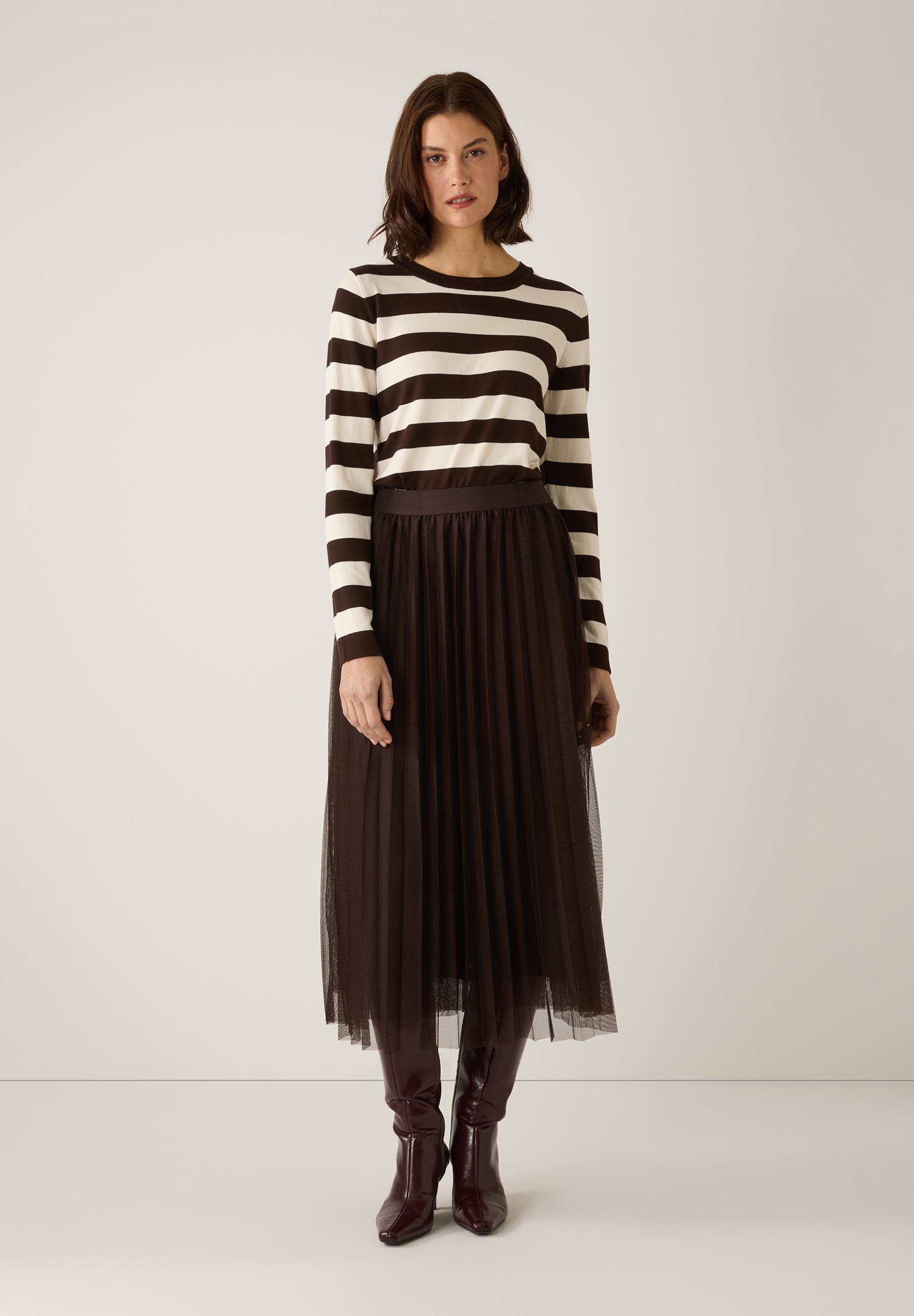 Eine Frau trägt einen MORE & MORE-Pullover mit braunen und weißen Streifen, einen Rock und Stiefel. Eine Frau trägt einen MORE & MORE-Pullover mit braunen und weißen Streifen, einen Rock und Stiefel.