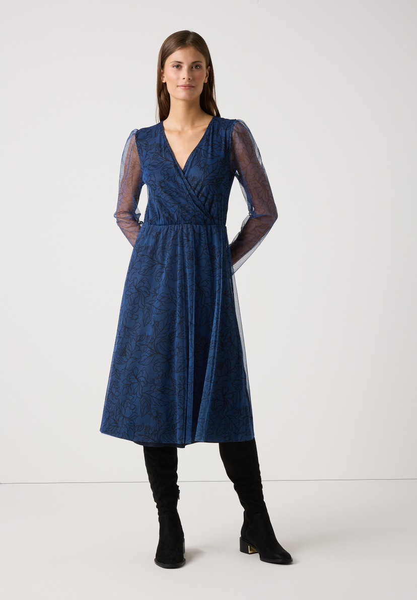 Eine Frau in einem blauen MORE & MORE Mesh-Kleid mit Blumenmuster und langen Ärmeln. Eine Frau in einem blauen MORE & MORE Mesh-Kleid mit Blumenmuster und langen Ärmeln.