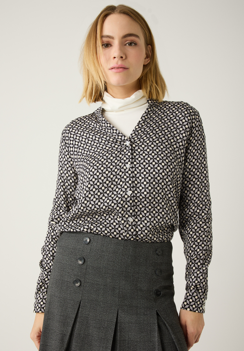 Eine Frau in MORE & MORE Oberteil mit Muster und grauem Rock mit Knöpfen vor hellem Hintergrund. Eine Frau in MORE & MORE Oberteil mit Muster und grauem Rock mit Knöpfen vor hellem Hintergrund.