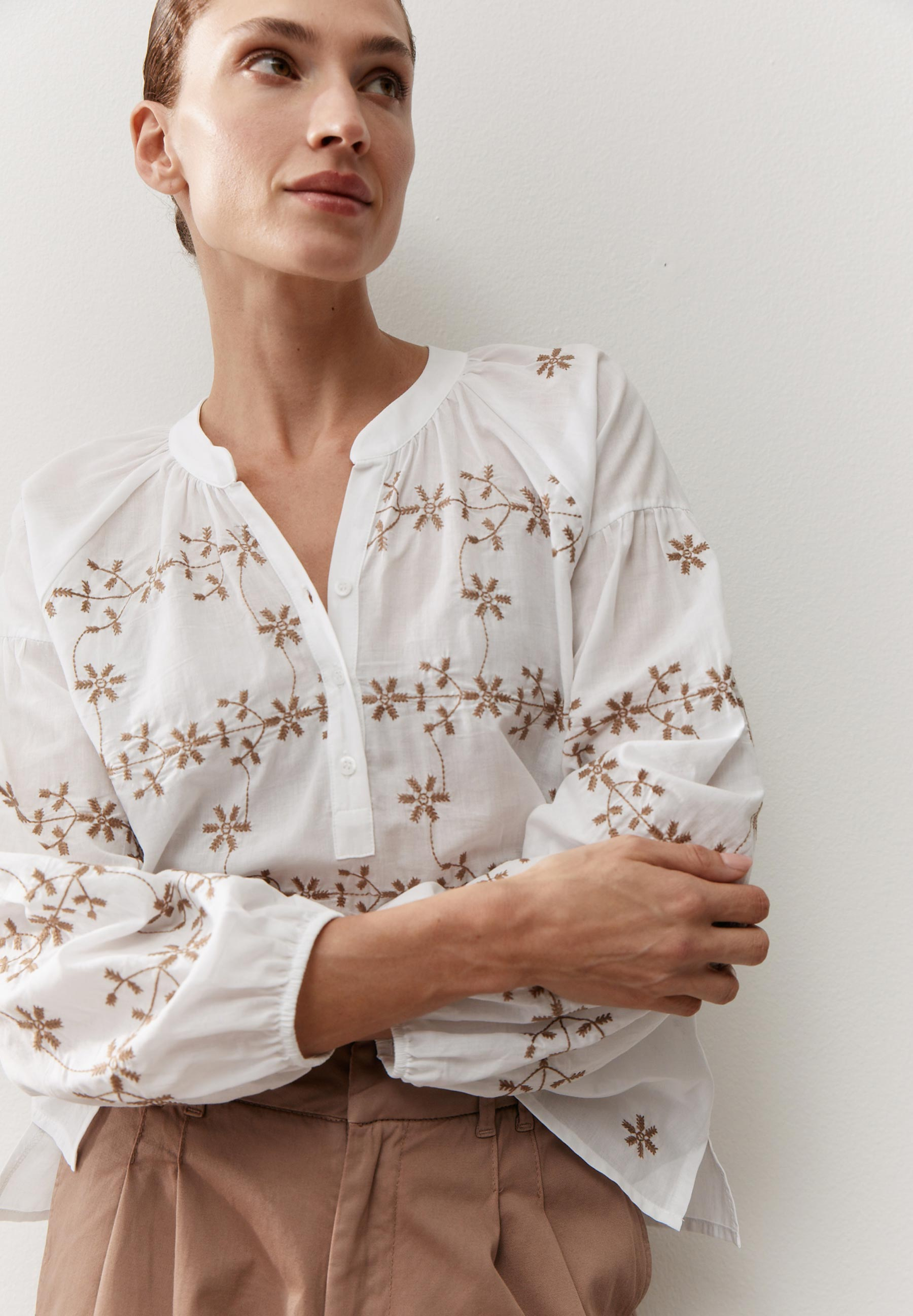 Eine MORE & MORE Bluse: Weißes, langärmeliges Oberteil mit braunen Stickereien, leicht geöffneter Kragen und zwei Knöpfe.