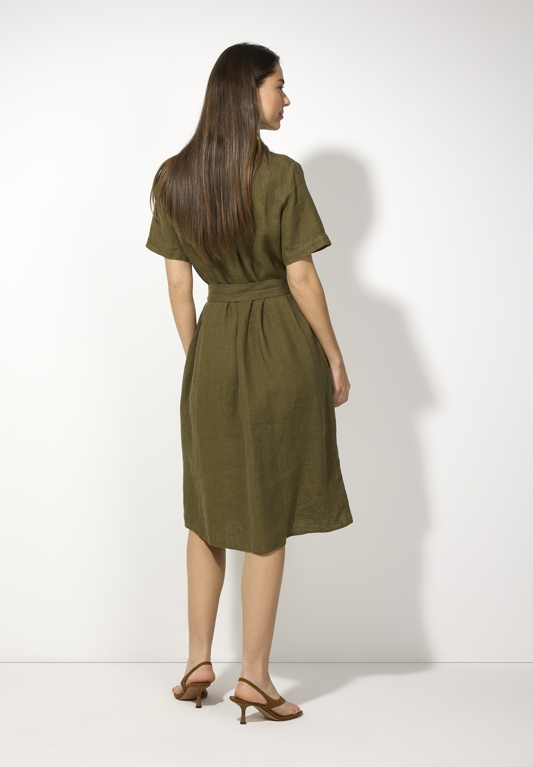 Eine Frau von hinten in einem olivfarbenen MORE & MORE Leinenkleid mit Gürtel und braunen Schuhen vor einem weißen Hintergrund. Eine Frau von hinten in einem olivfarbenen MORE & MORE Leinenkleid mit Gürtel und braunen Schuhen vor einem weißen Hintergrund.