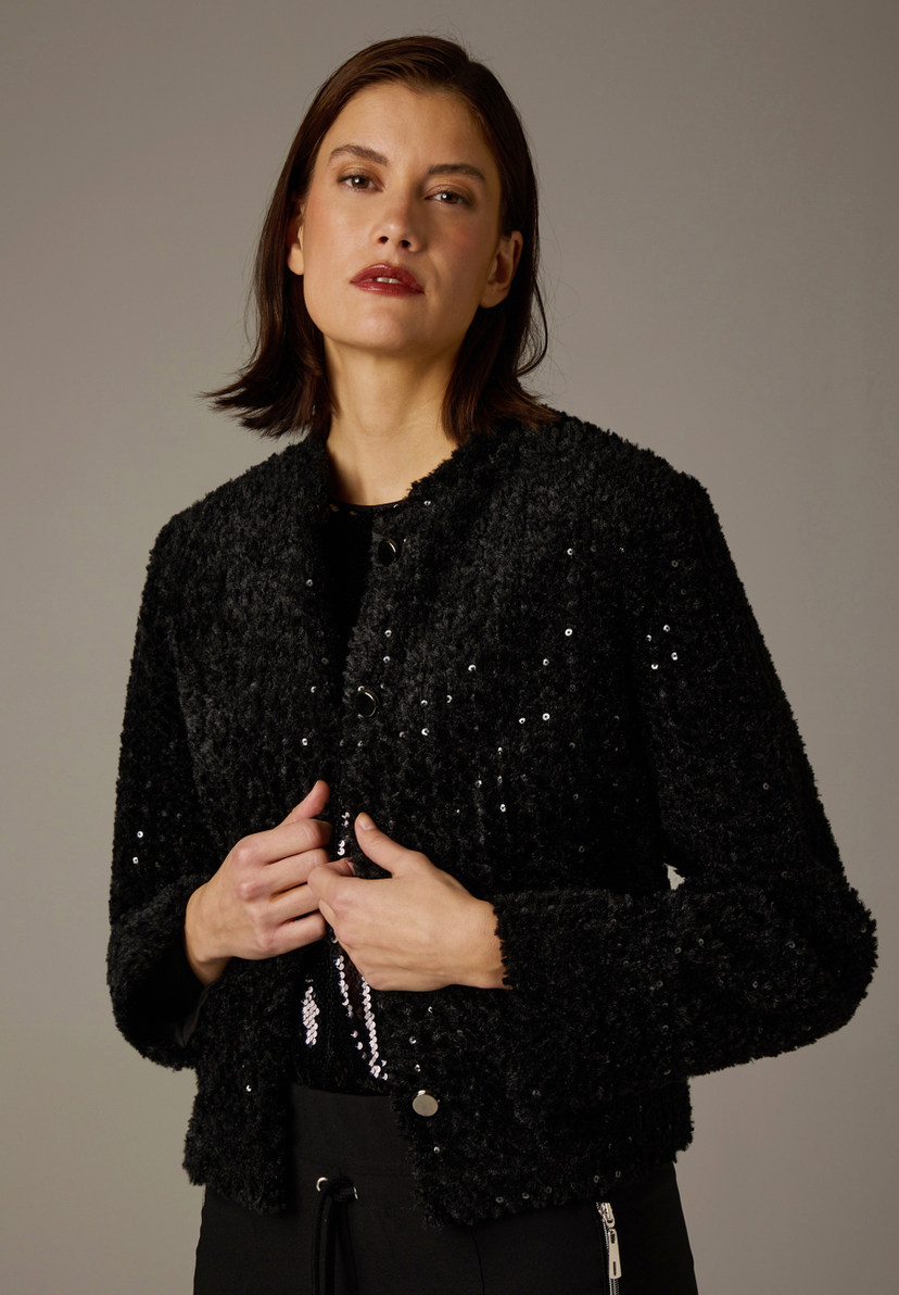 Eine Frau präsentiert einen schwarzen MORE & MORE Blazer mit glitzernden Pailletten. Sie hält ihn mit den Händen leicht geschlossen. Eine Frau präsentiert einen schwarzen MORE & MORE Blazer mit glitzernden Pailletten. Sie hält ihn mit den Händen leicht geschlossen.