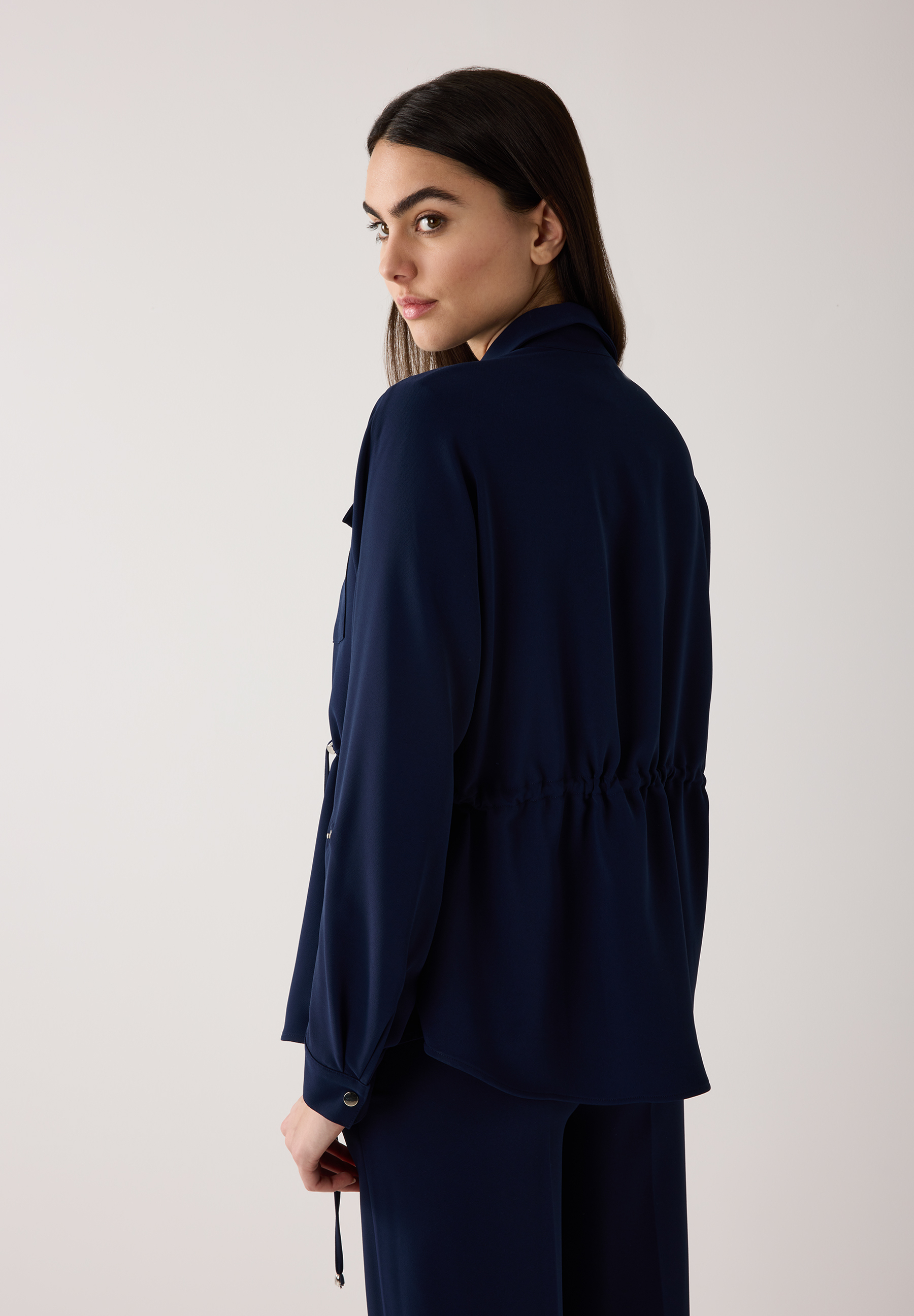 Eine Frau blickt über ihre Schulter und trägt ein marineblaues Blazer-Oberteil von MORE & MORE mit Webware-Konstruktion. Eine Frau blickt über ihre Schulter und trägt ein marineblaues Blazer-Oberteil von MORE & MORE mit Webware-Konstruktion.