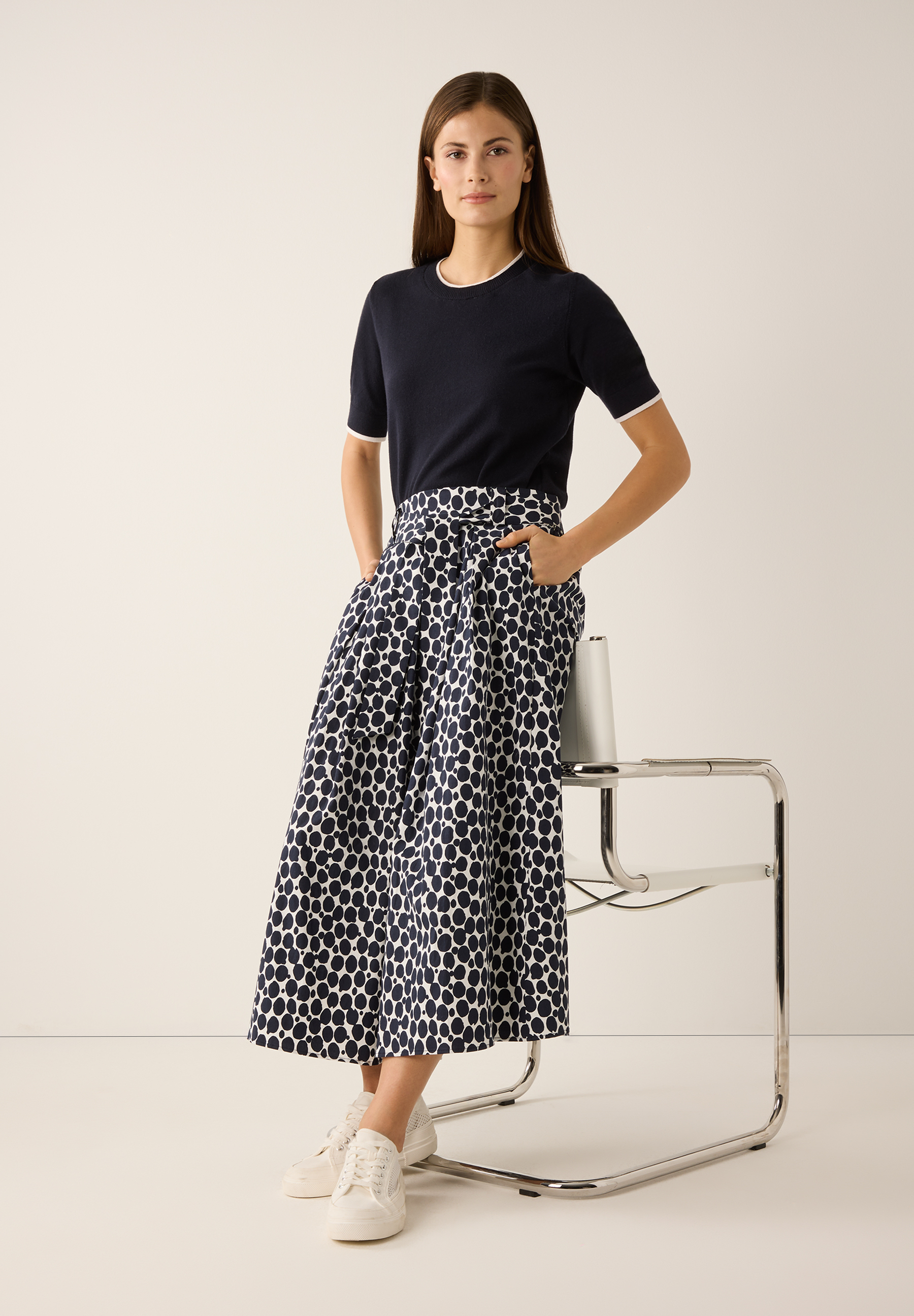 Eine Frau mit einem MORE & MORE Feinstrick-Oberteil und einem Rock mit Punktmuster posiert lässig an einem Stuhl. Eine Frau mit einem MORE & MORE Feinstrick-Oberteil und einem Rock mit Punktmuster posiert lässig an einem Stuhl.