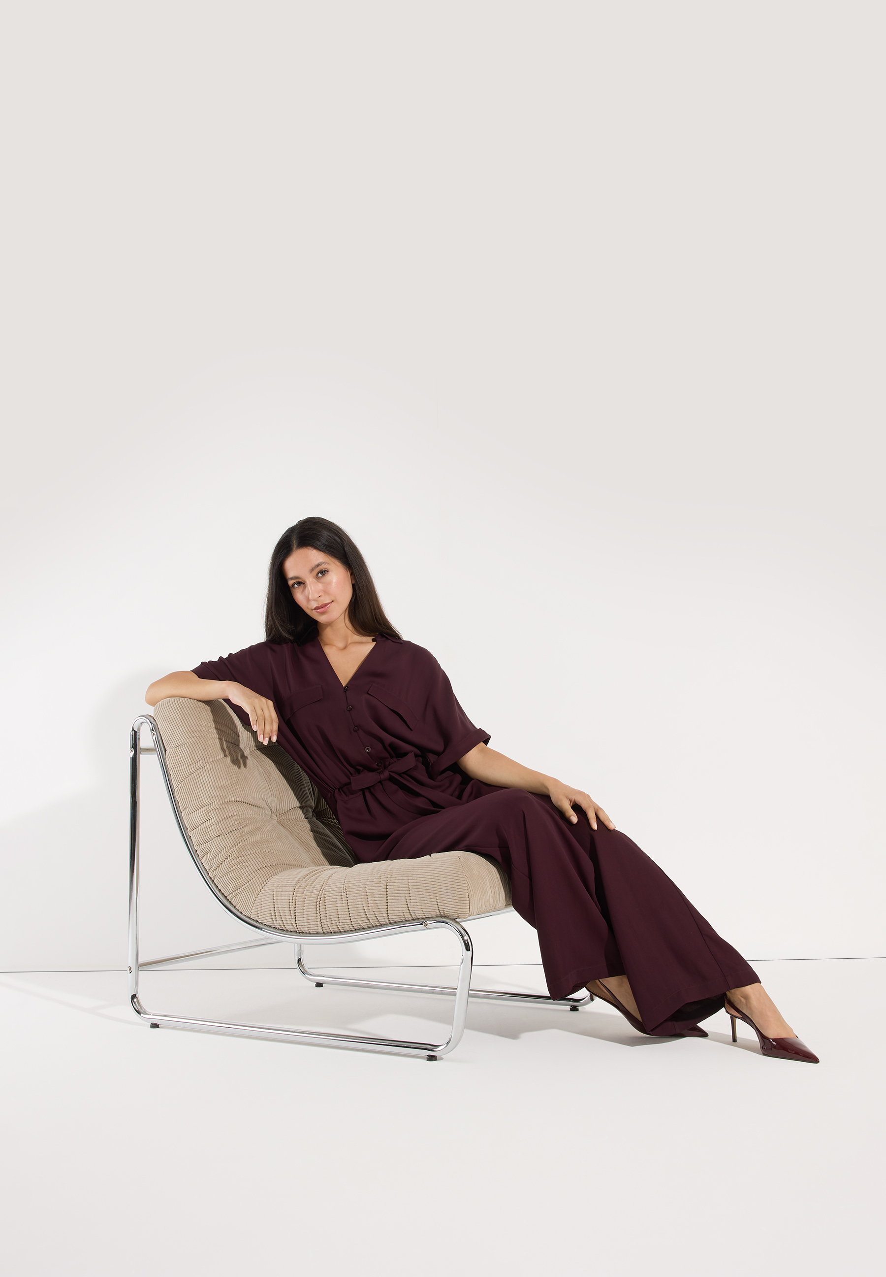 MORE & MORE Jeans: Stilvolle Frau im bordeauxroten Jumpsuit posiert elegant auf einem Sessel vor weißem Hintergrund. MORE & MORE Jeans: Stilvolle Frau im bordeauxroten Jumpsuit posiert elegant auf einem Sessel vor weißem Hintergrund.