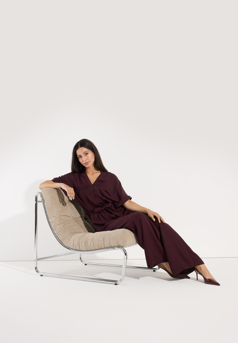 MORE & MORE Jeans: Stilvolle Frau im bordeauxroten Jumpsuit posiert elegant auf einem Sessel vor weißem Hintergrund. MORE & MORE Jeans: Stilvolle Frau im bordeauxroten Jumpsuit posiert elegant auf einem Sessel vor weißem Hintergrund.