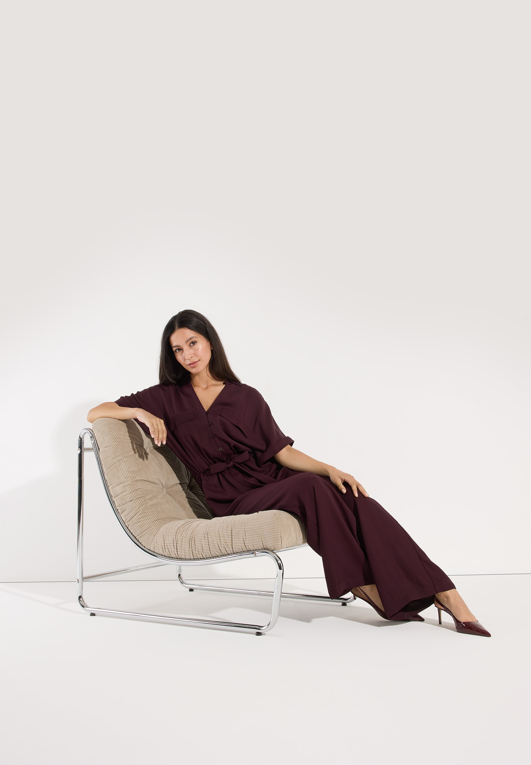 MORE & MORE Jeans: Stilvolle Frau im bordeauxroten Jumpsuit posiert elegant auf einem Sessel vor weißem Hintergrund. MORE & MORE Jeans: Stilvolle Frau im bordeauxroten Jumpsuit posiert elegant auf einem Sessel vor weißem Hintergrund.