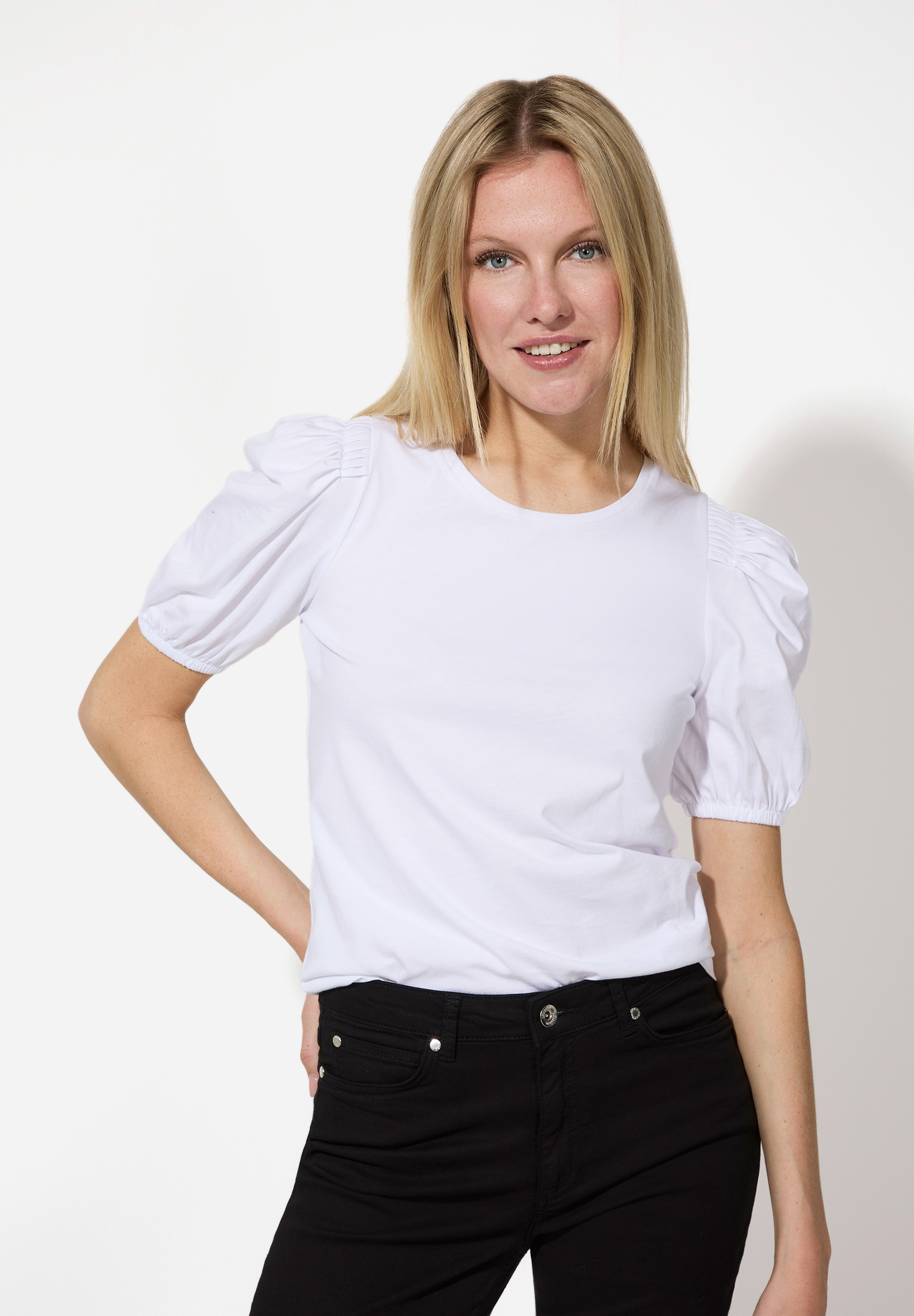 Eine Frau im MORE & MORE Jersey-Top mit Puffärmeln und schwarzen Jeans vor einem weißen Hintergrund.