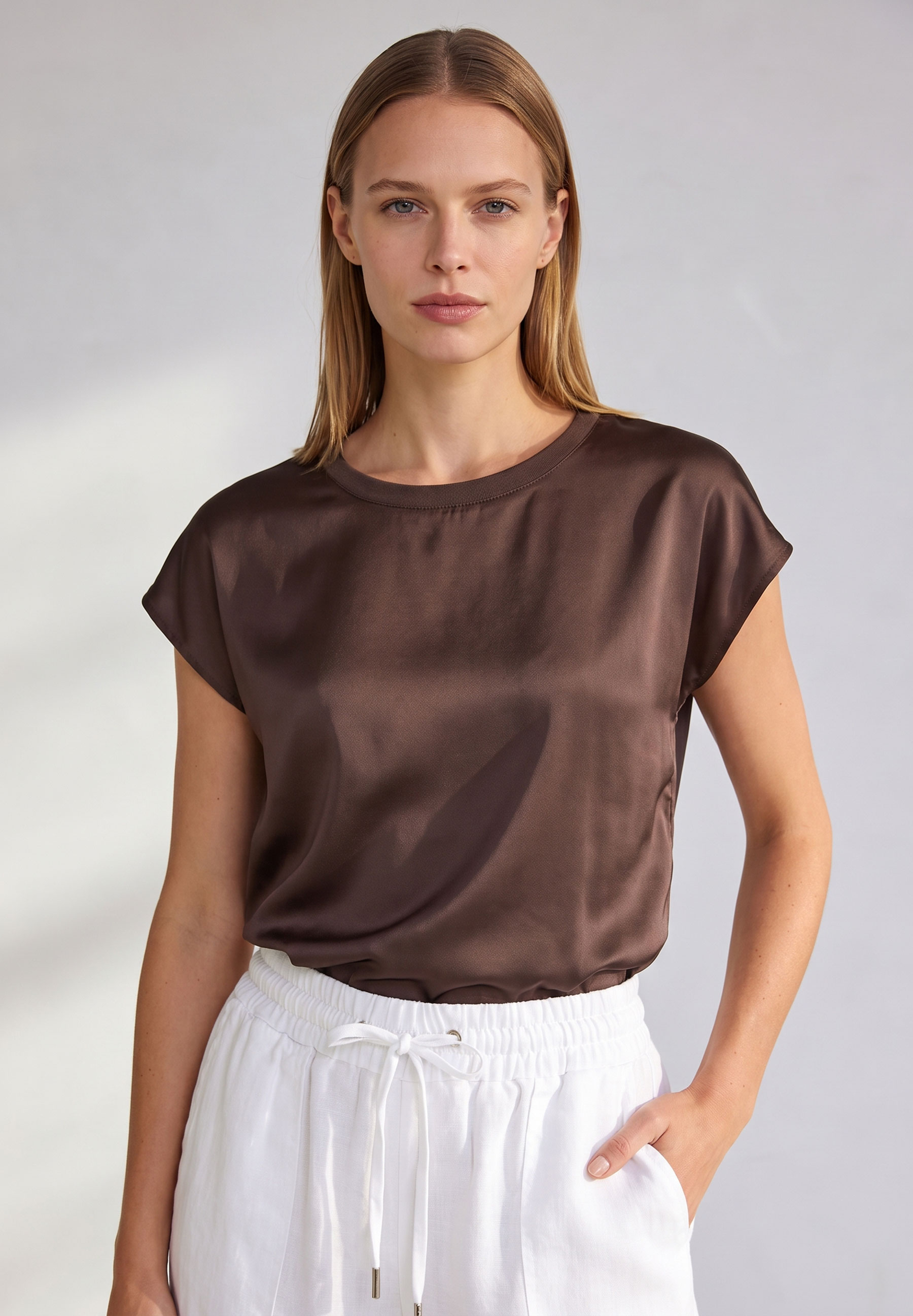 AI generated - MORE & MORE T-Shirt aus Satin in Braun, kombiniert mit weißen Shorts; die Frau blickt frontal in die Kamera.