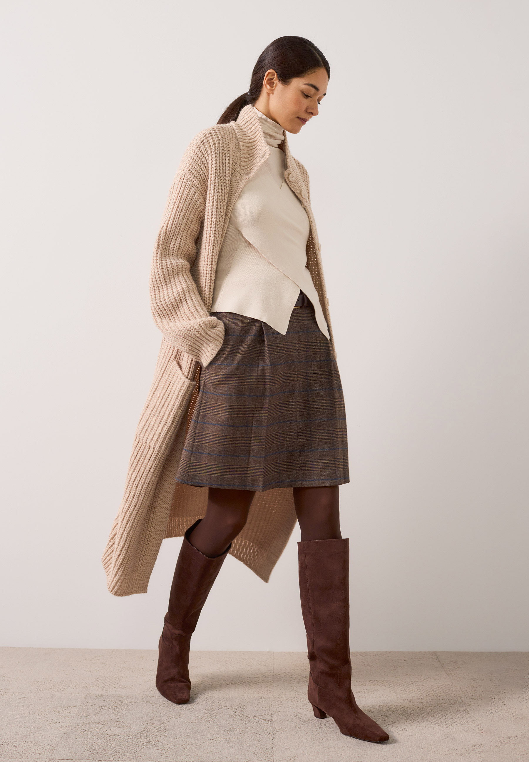 MORE & MORE langer Strickpullover in Beige, dazu karierter Rock, Rollkragenpullover und braune Stiefel.