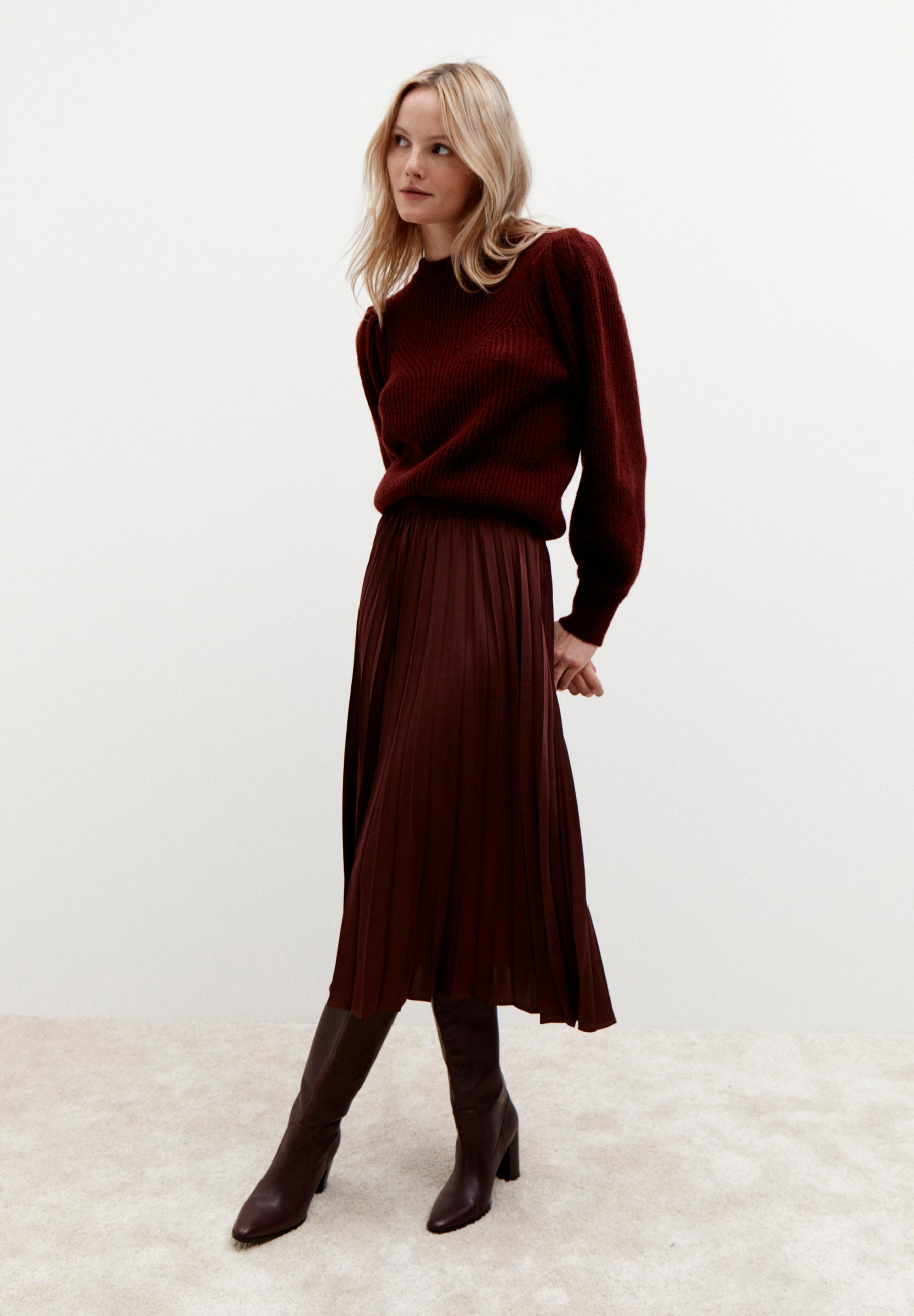 Eine blonde Frau trägt einen MORE & MORE Strickpullover in Burgunderrot, einen Faltenrock und passende Stiefel vor einem weißen Hintergrund. Eine blonde Frau trägt einen MORE & MORE Strickpullover in Burgunderrot, einen Faltenrock und passende Stiefel vor einem weißen Hintergrund.