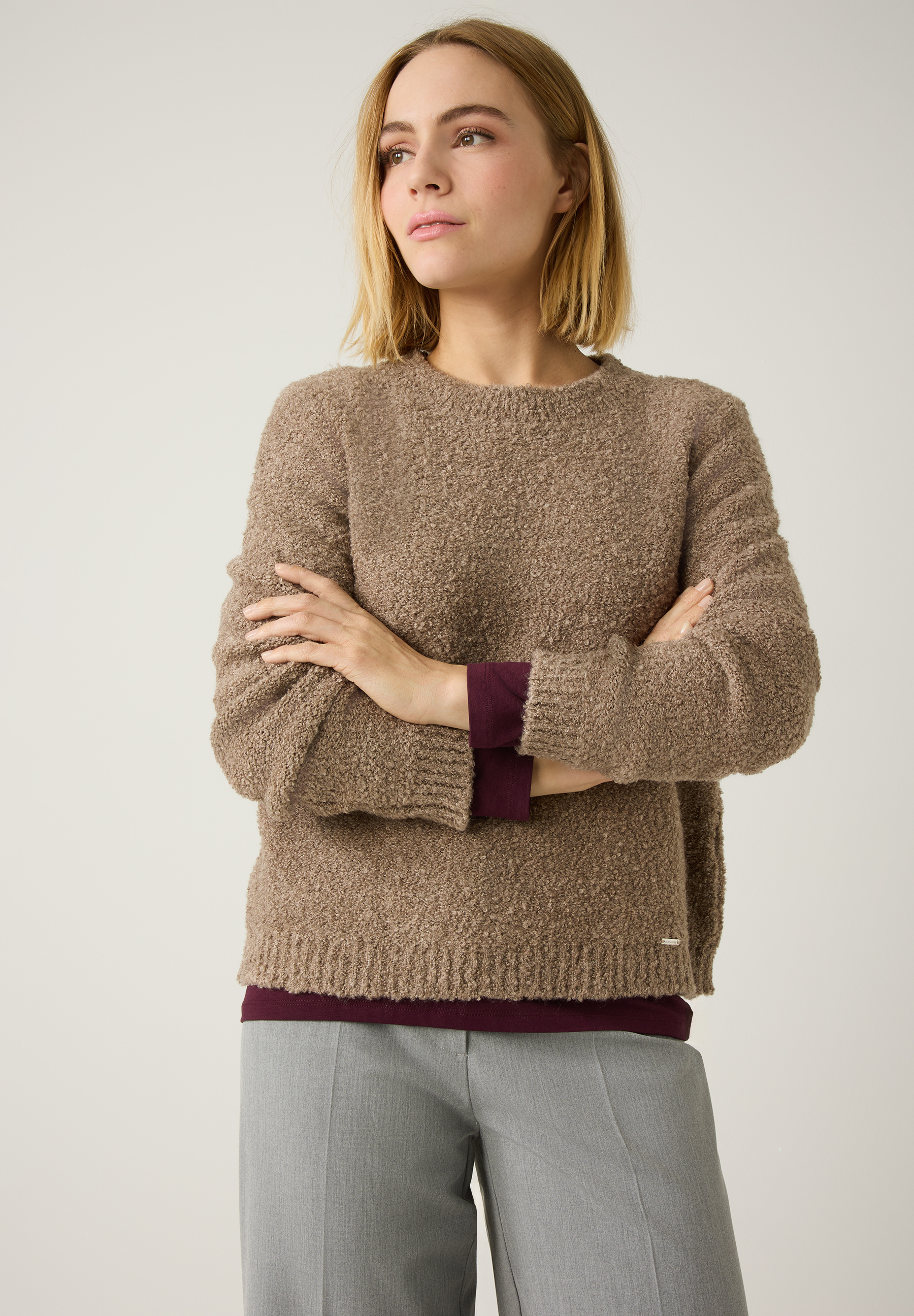 Eine Frau mit blonden Haaren trägt einen beigen Strickpullover der Marke MORE & MORE und eine graue Hose. Sie hat die Arme verschränkt.