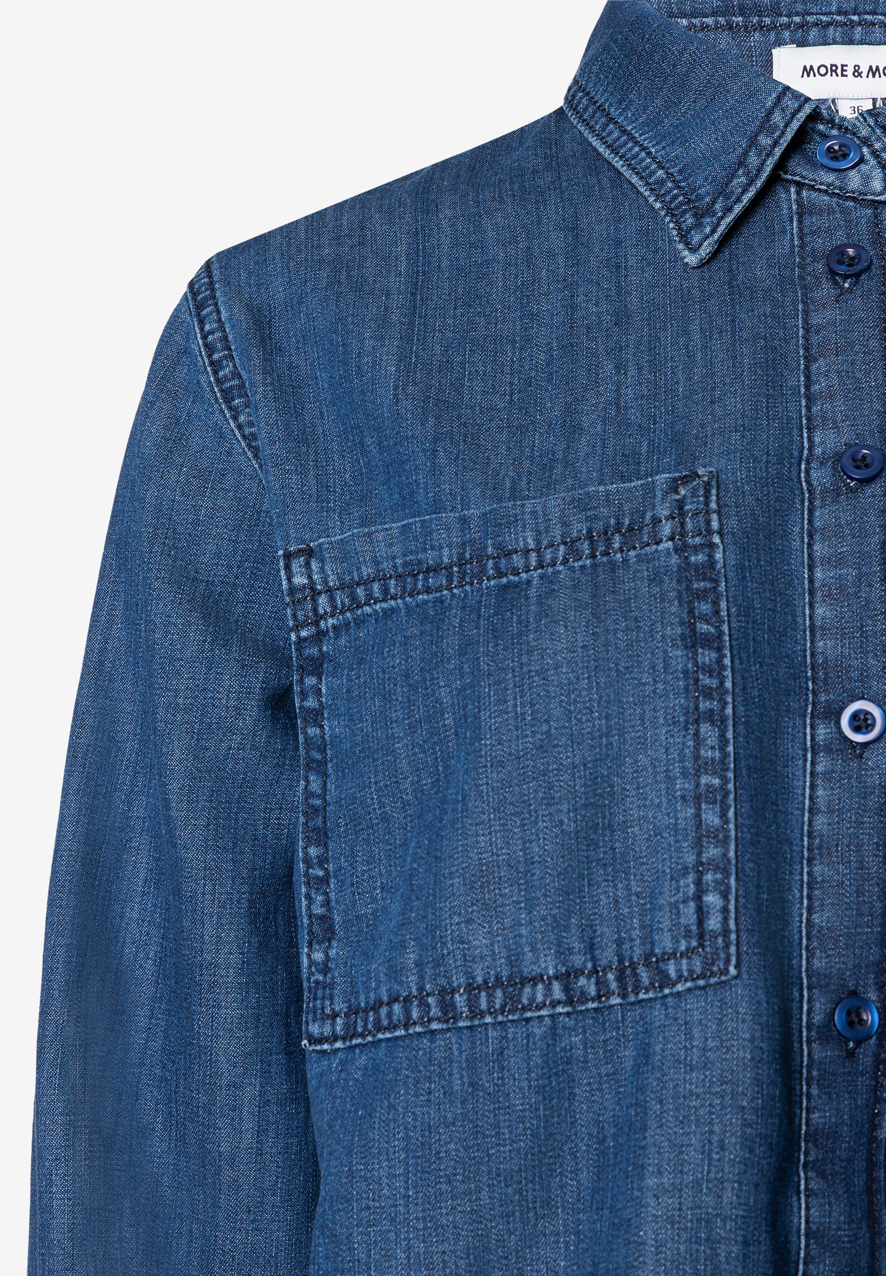 Detailaufnahme einer MORE & MORE Bluse aus Denim mit Brusttasche und blauen Knöpfen. Angenehmer Tragekomfort. Detailaufnahme einer MORE & MORE Bluse aus Denim mit Brusttasche und blauen Knöpfen. Angenehmer Tragekomfort.