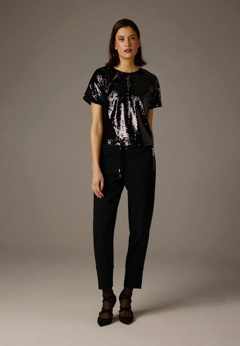 Eine Frau in MORE & MORE Hose und Pailletten-Shirt, posiert vor einem grauen Hintergrund. Eine Frau in MORE & MORE Hose und Pailletten-Shirt, posiert vor einem grauen Hintergrund.