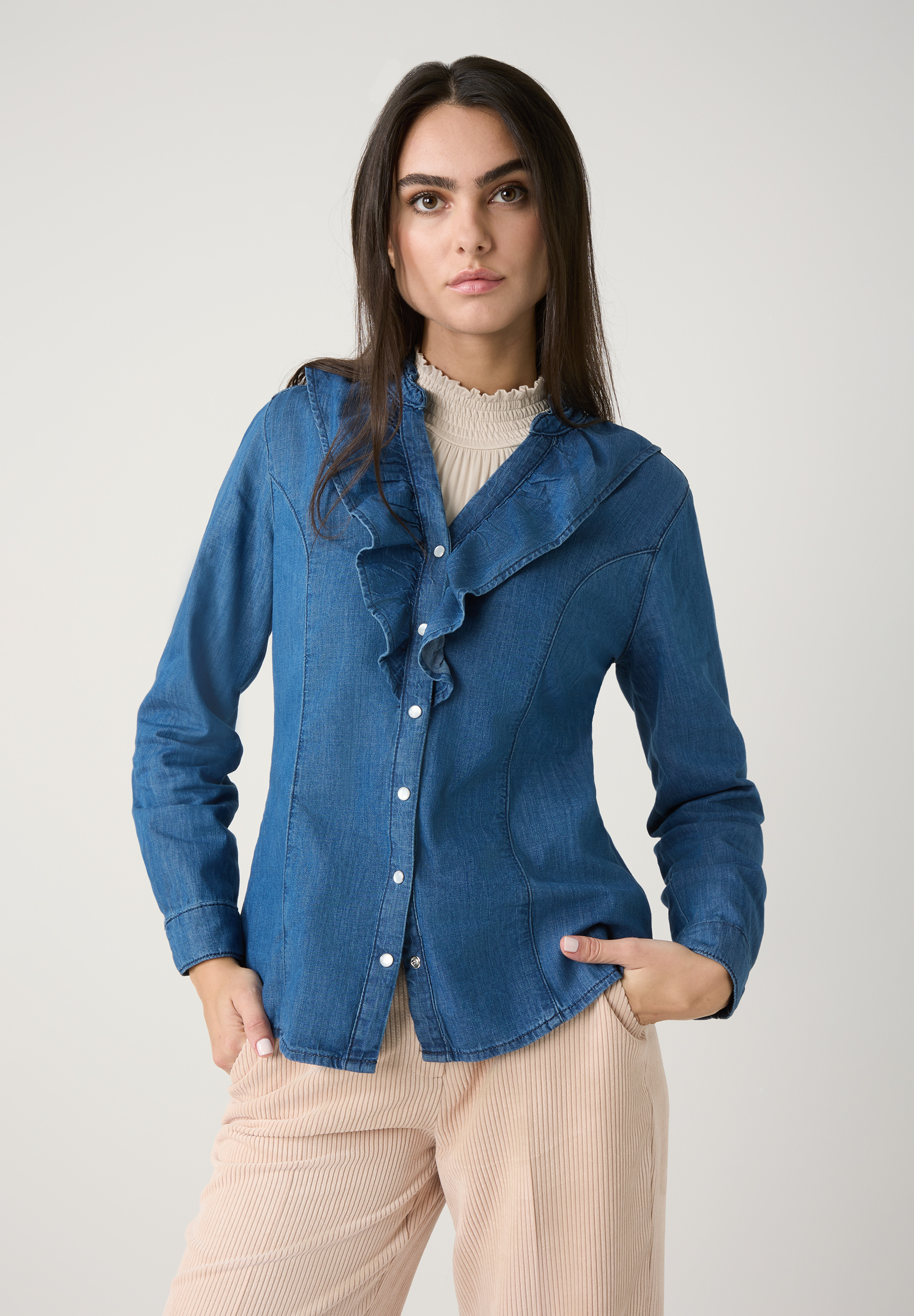 Eine Frau in einer blauen MORE & MORE Bluse mit Rüschen und beigen Hosen posiert vor einem neutralen Hintergrund.