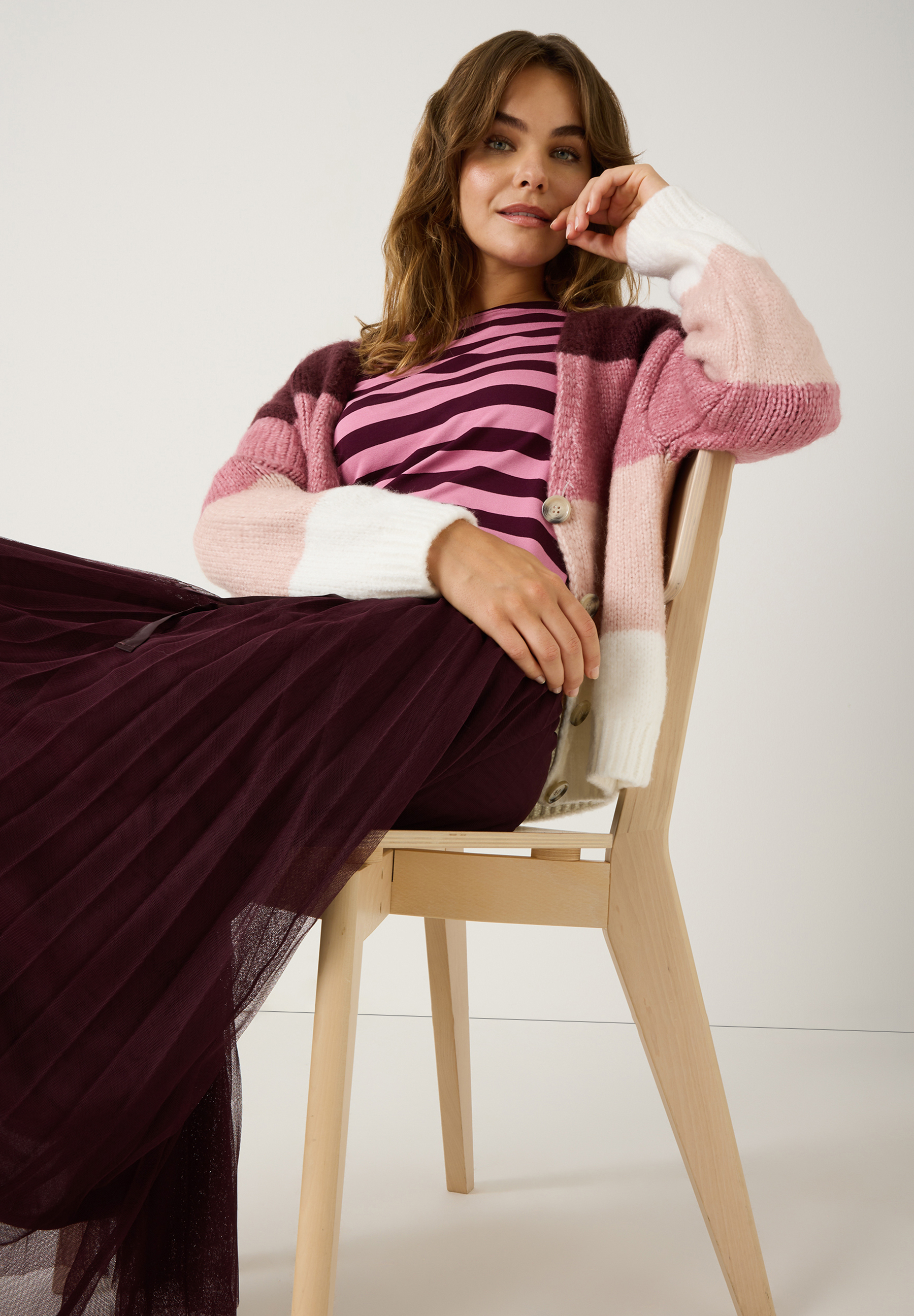 Eine Frau sitzt auf einem Stuhl und trägt MORE & MORE Strick-Oberteile in Rosa- und Lilatönen. Eine Frau sitzt auf einem Stuhl und trägt MORE & MORE Strick-Oberteile in Rosa- und Lilatönen.