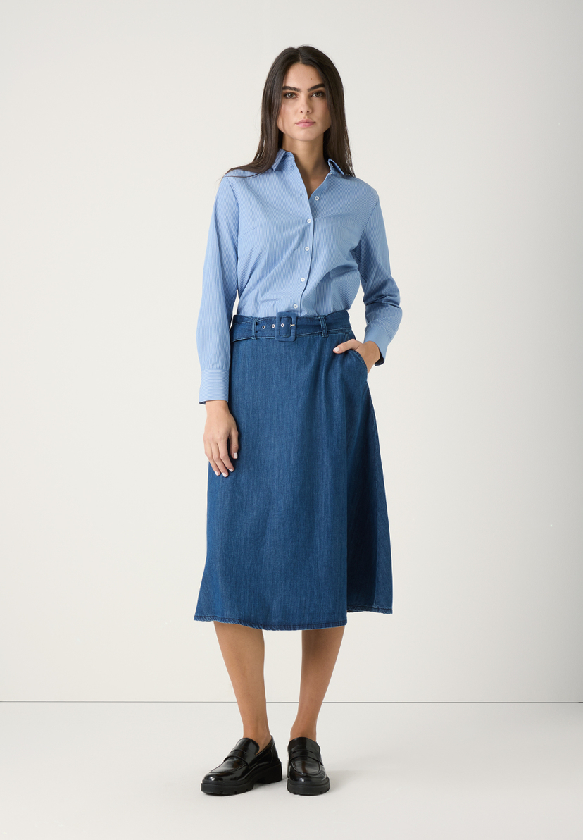 Eine Frau mit Rock von MORE & MORE aus Denim mit angenehmem Tragekomfort und blau gestreifter Bluse, stehend vor hellem Hintergrund. Eine Frau mit Rock von MORE & MORE aus Denim mit angenehmem Tragekomfort und blau gestreifter Bluse, stehend vor hellem Hintergrund.