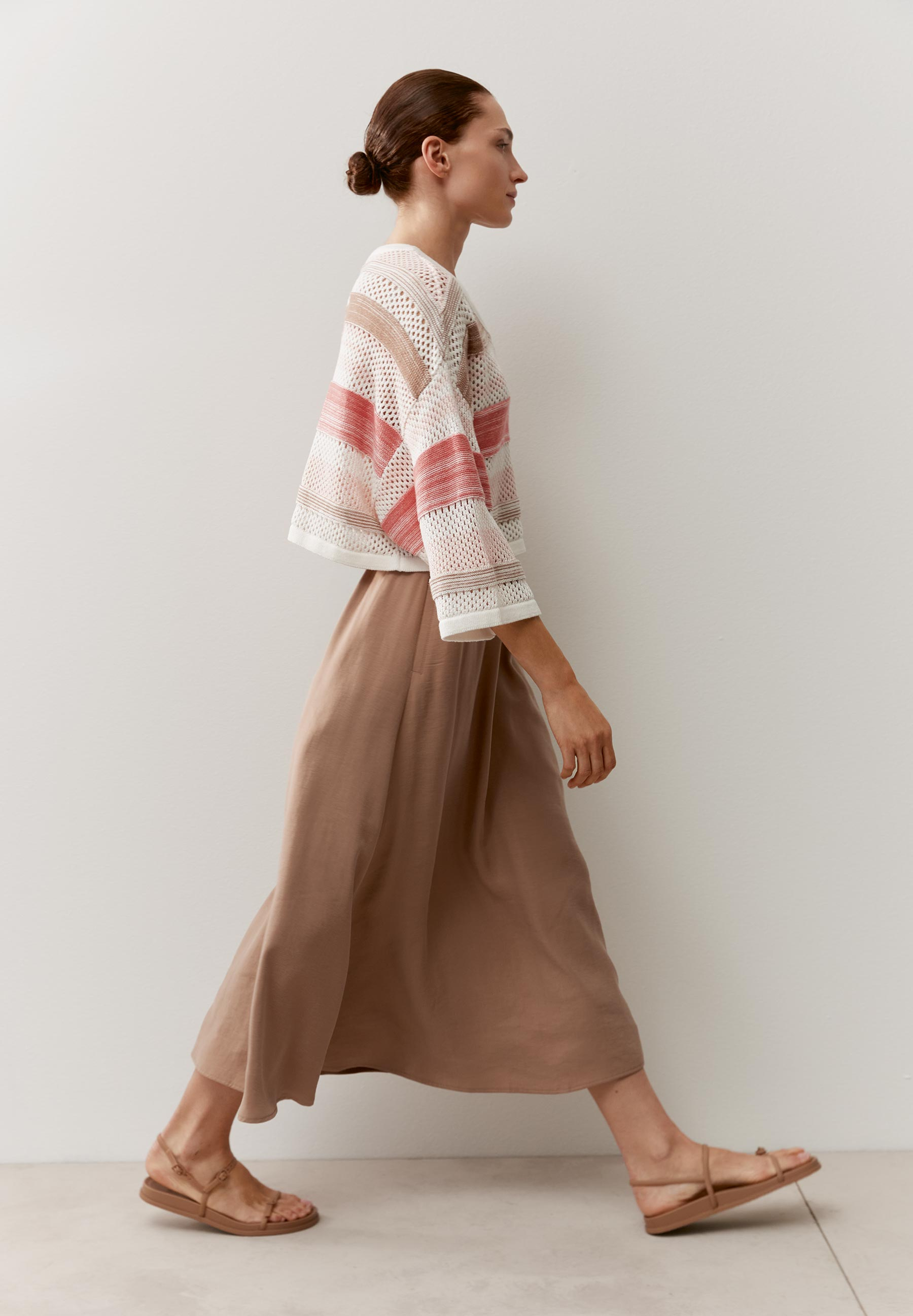 Eine Frau in MORE & MORE Strickware mit Streifen, dazu ein langer Rock und Sandalen. Sie geht vor einer weißen Wand entlang.