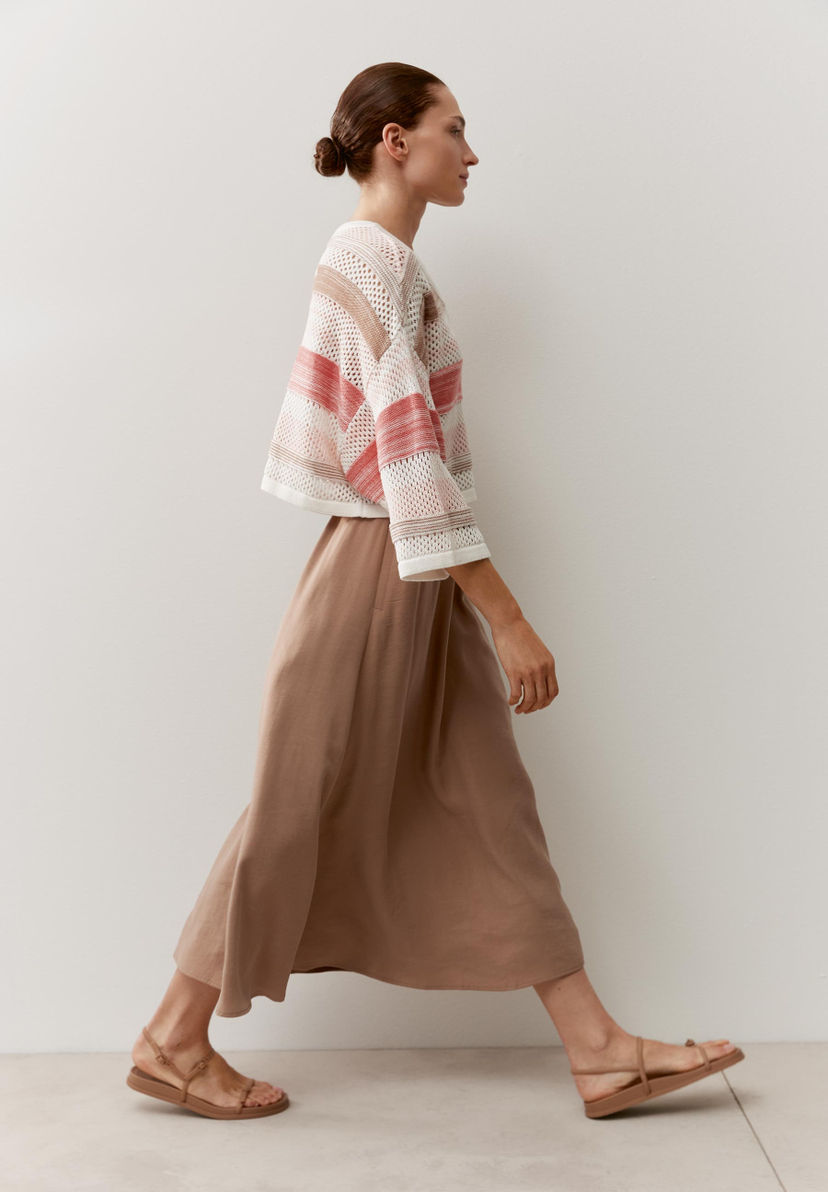 Eine Frau in MORE & MORE Strickware mit Streifen, dazu ein langer Rock und Sandalen. Sie geht vor einer weißen Wand entlang.