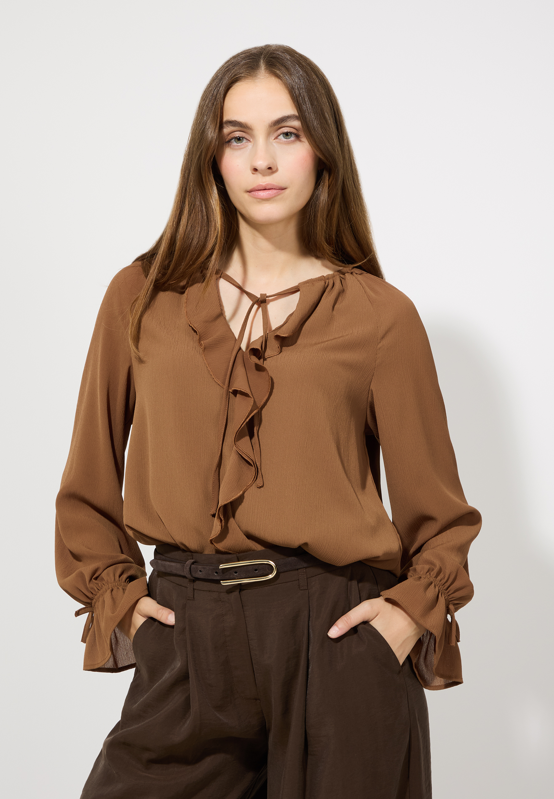 Eine Frau mit braunen MORE & MORE Gürtel posiert in brauner Bluse und Hose vor einem hellen Hintergrund.
