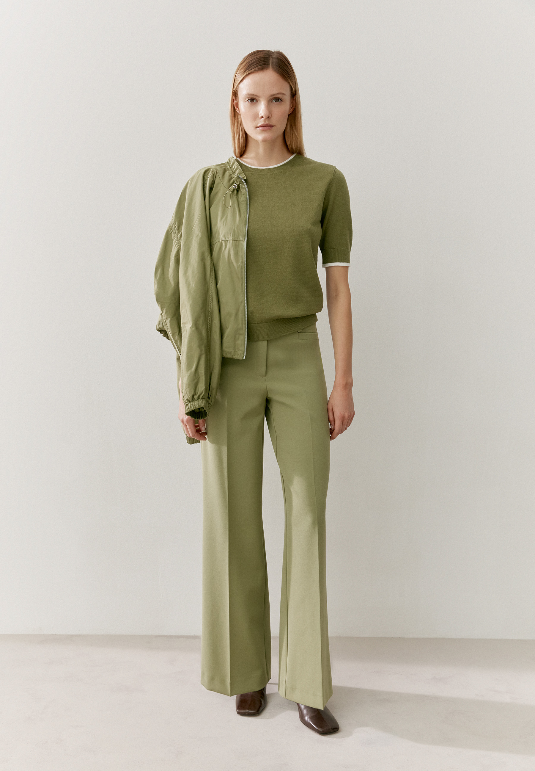 Eine Frau trägt ein olivgrünes MORE & MORE Feinstrick-Oberteil mit passender Jacke und Hose vor einer weißen Wand. Eine Frau trägt ein olivgrünes MORE & MORE Feinstrick-Oberteil mit passender Jacke und Hose vor einer weißen Wand.