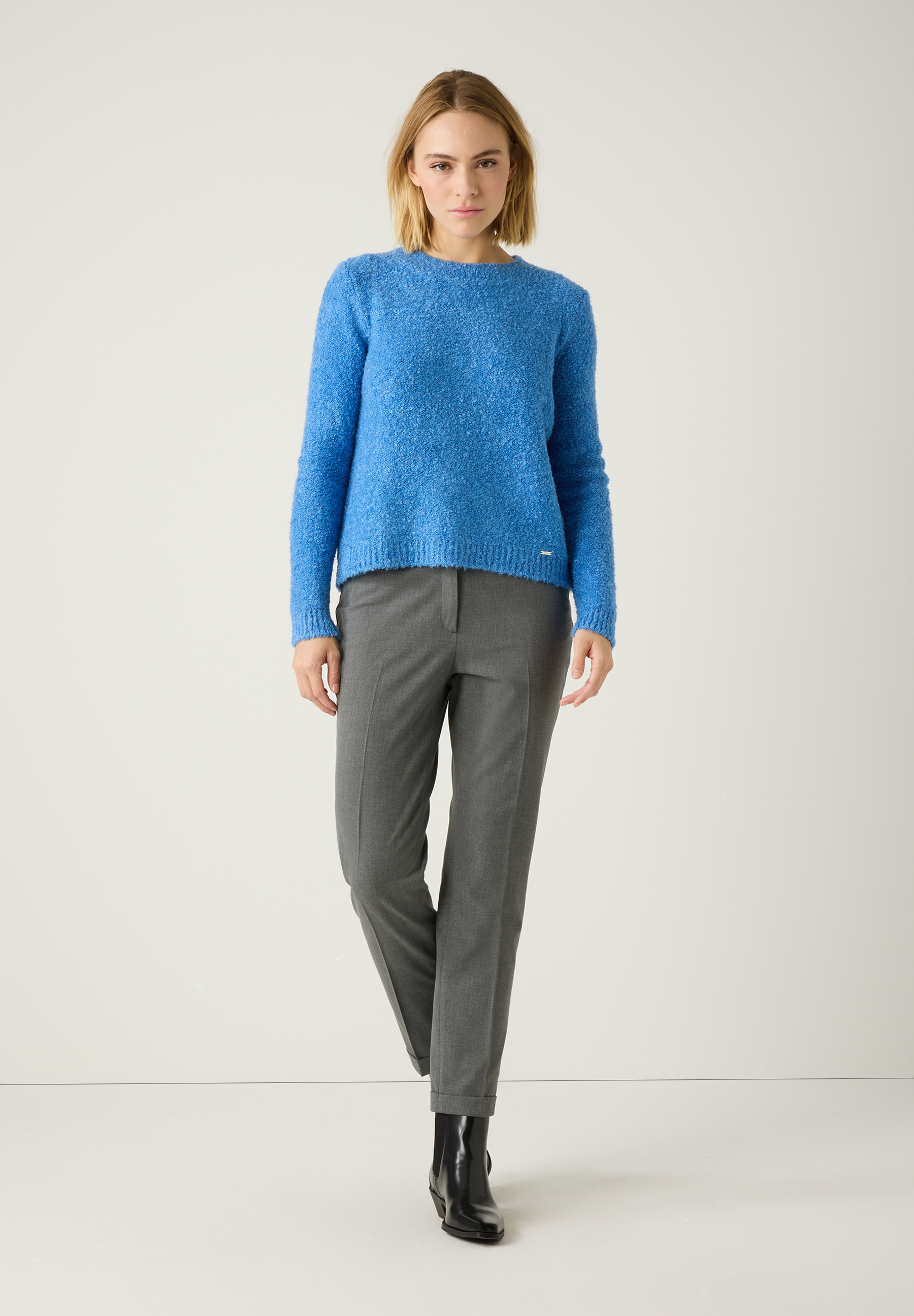 Eine Frau trägt einen blauen MORE & MORE Bouclé-Pullover zu grauer Hose und schwarzen Stiefeln. Eine Frau trägt einen blauen MORE & MORE Bouclé-Pullover zu grauer Hose und schwarzen Stiefeln.