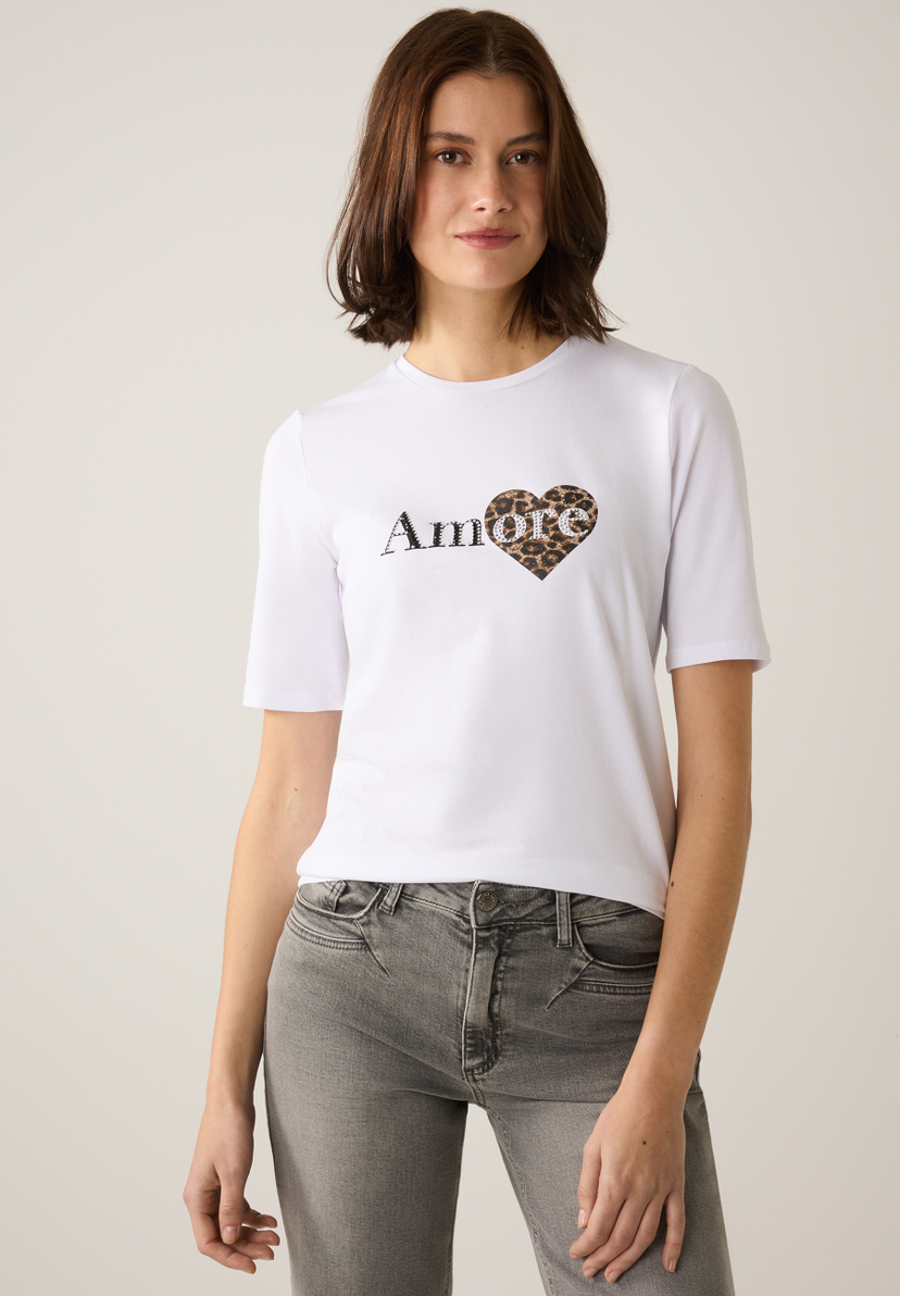 Eine Frau in einem weißen MORE & MORE T-Shirt mit „Amore“-Aufdruck und Leopardenherz. Eine Frau in einem weißen MORE & MORE T-Shirt mit „Amore“-Aufdruck und Leopardenherz.