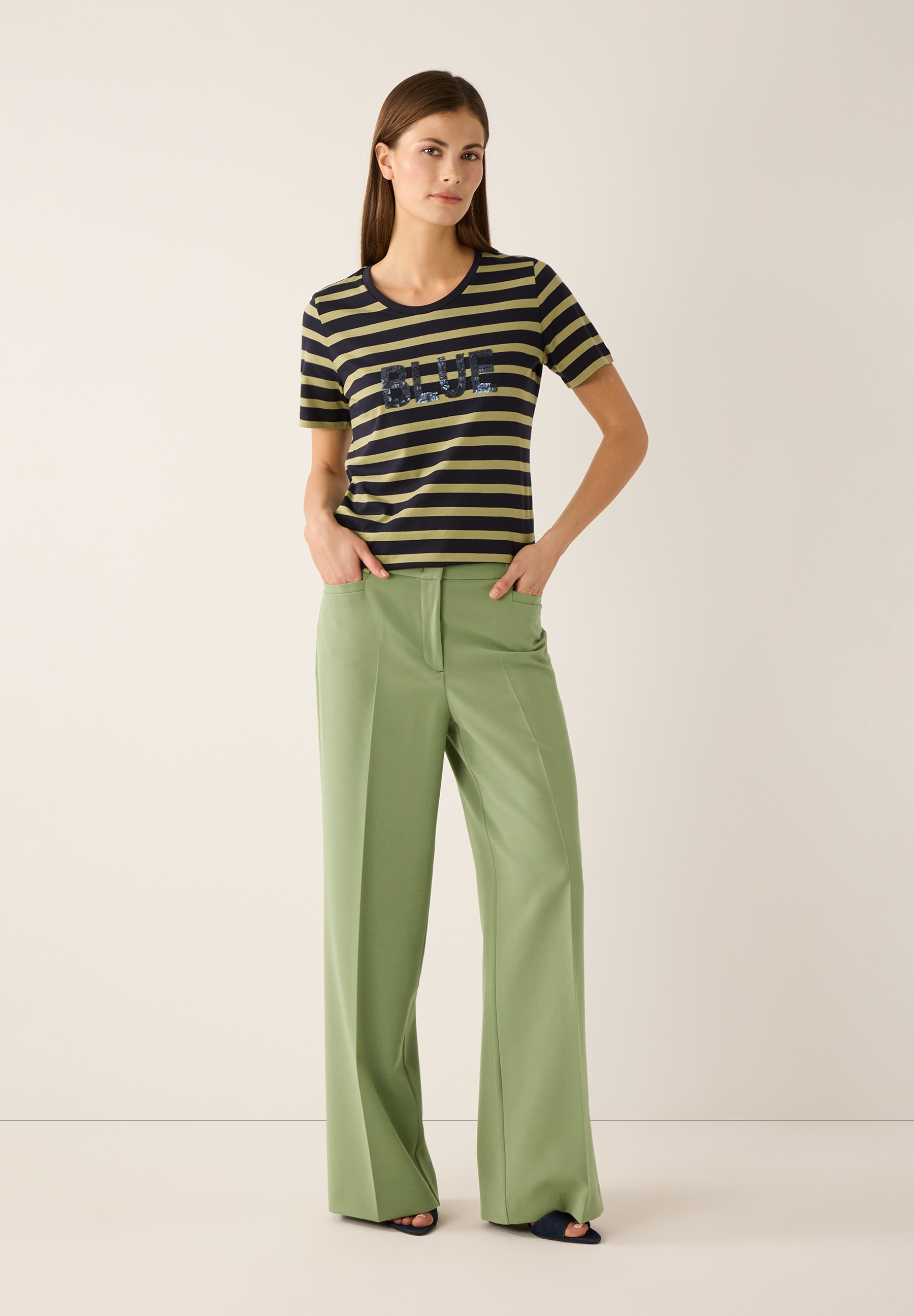 Eine Frau mit MORE & MORE Hose in Olivgrün mit weitem Bein und T-Shirt mit Streifenmuster. Eine Frau mit MORE & MORE Hose in Olivgrün mit weitem Bein und T-Shirt mit Streifenmuster.